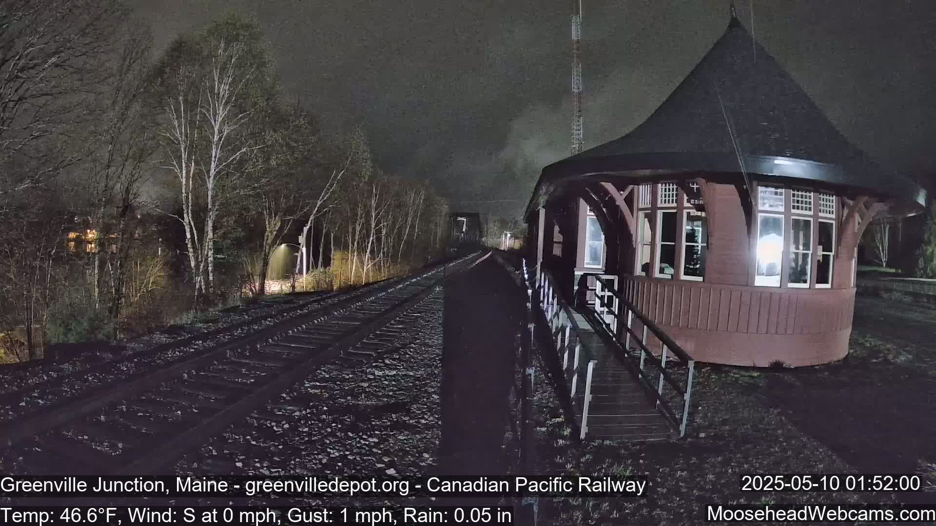 A dark red, round train depot sits beside railroad tracks under a nighttime rain.