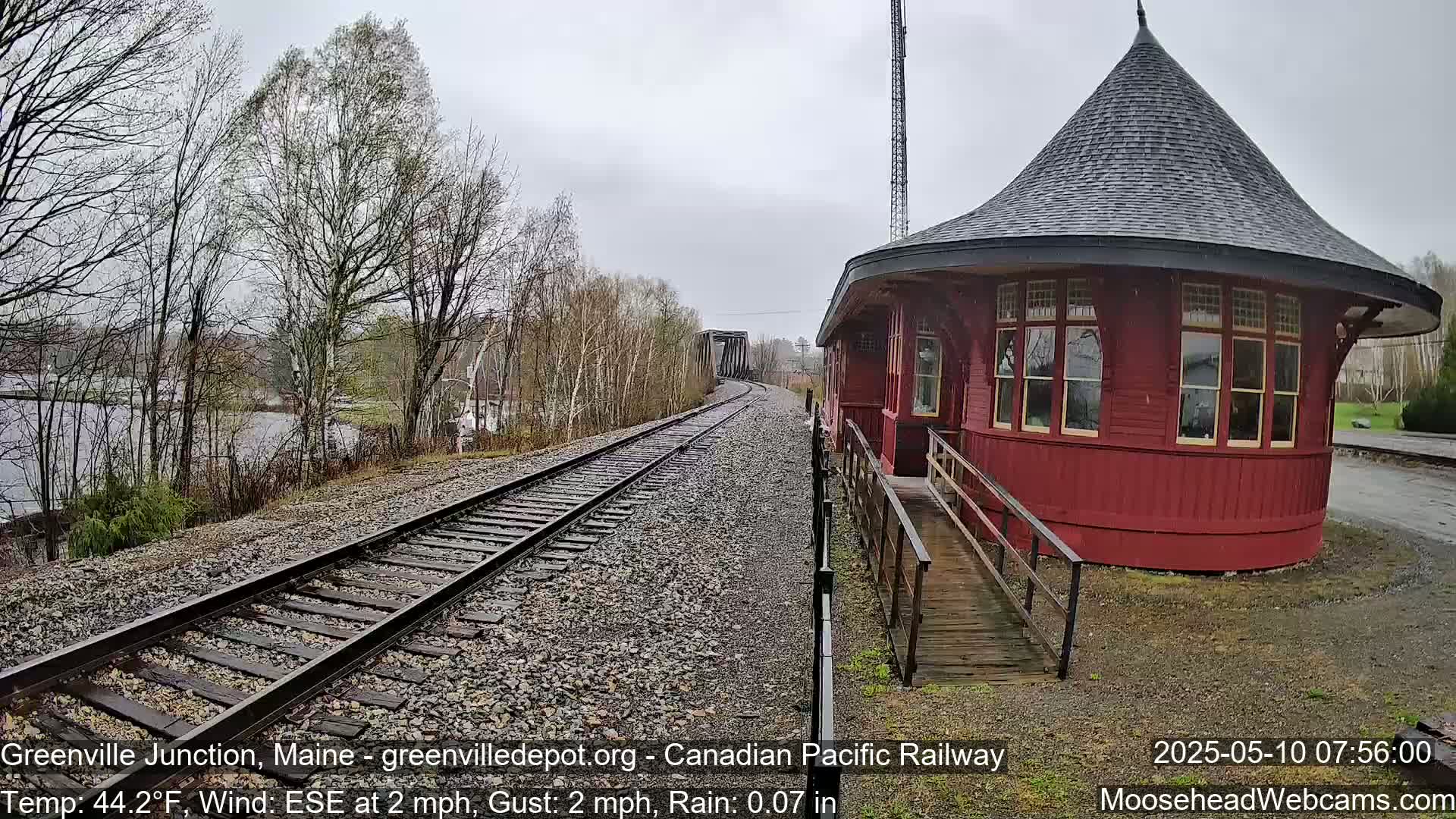 A red octagonal train depot with a conical roof sits beside railway tracks leading to a bridge, under a light rain.