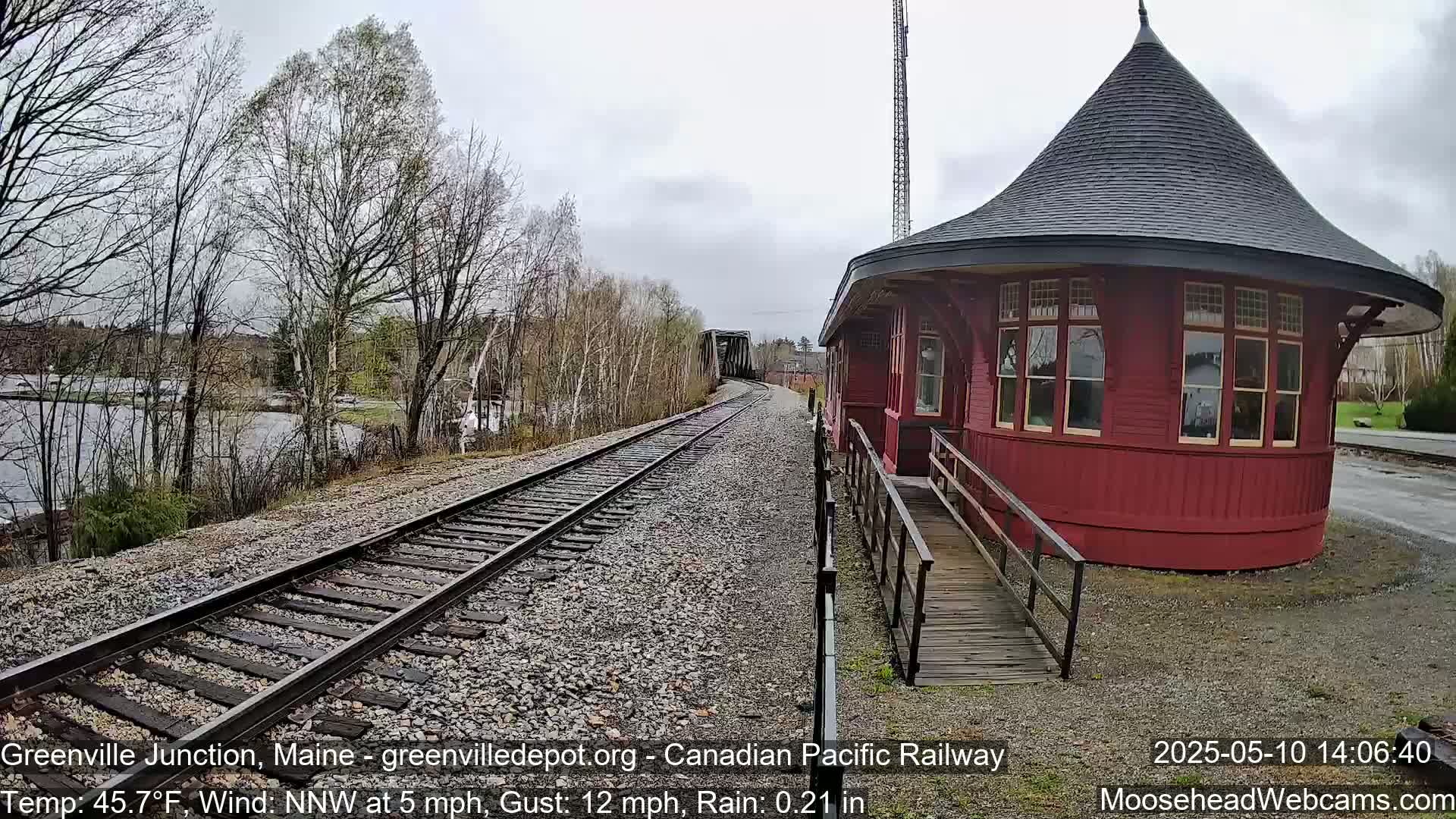 A red, round train depot with a conical roof sits beside railroad tracks leading to a bridge, under a light rain.