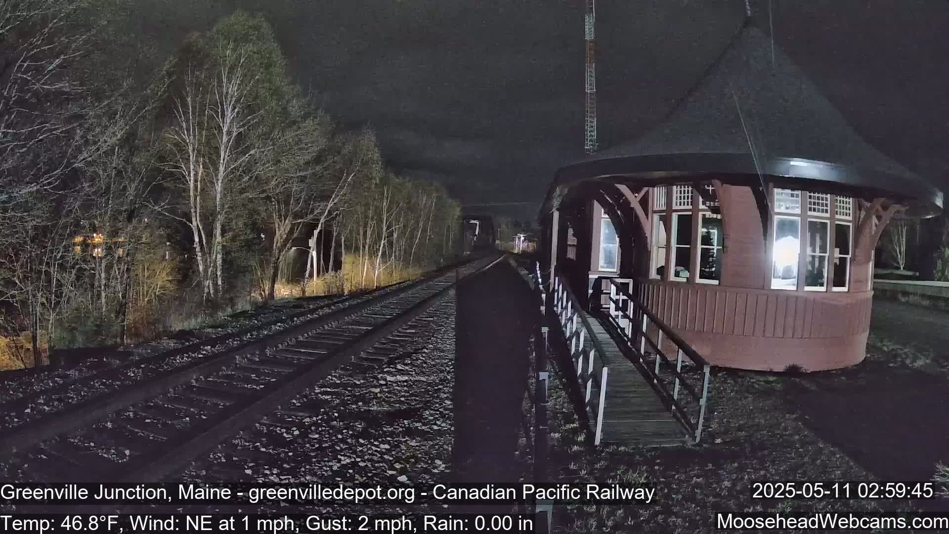 A dark-red, round train depot stands beside railroad tracks that extend into a dark, wooded area at night, with a northeast wind of 1 mph and gusts up to 2 mph.