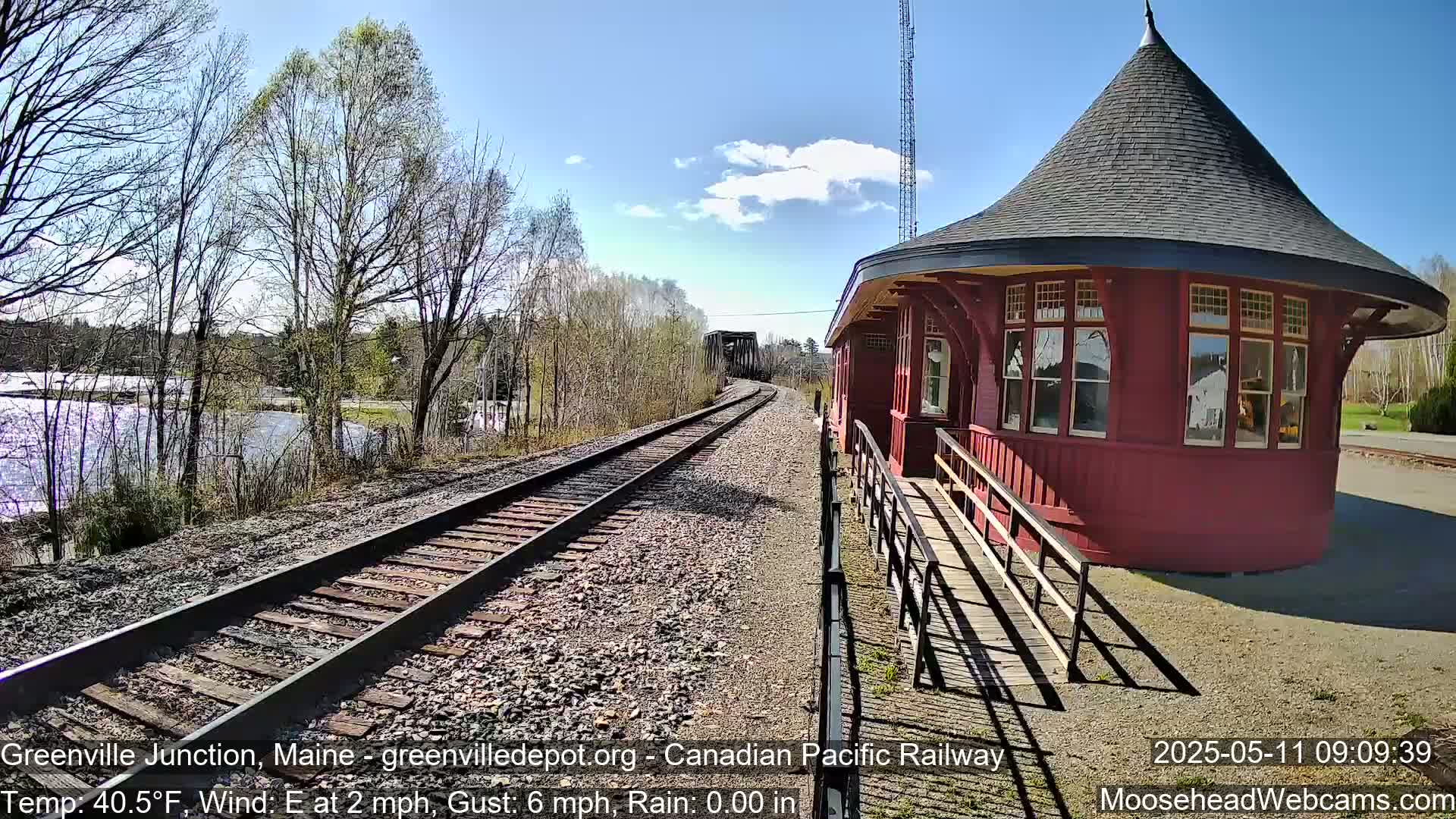 A red, octagonal train depot stands beside railroad tracks curving toward a bridge and a body of water under a partly cloudy sky.