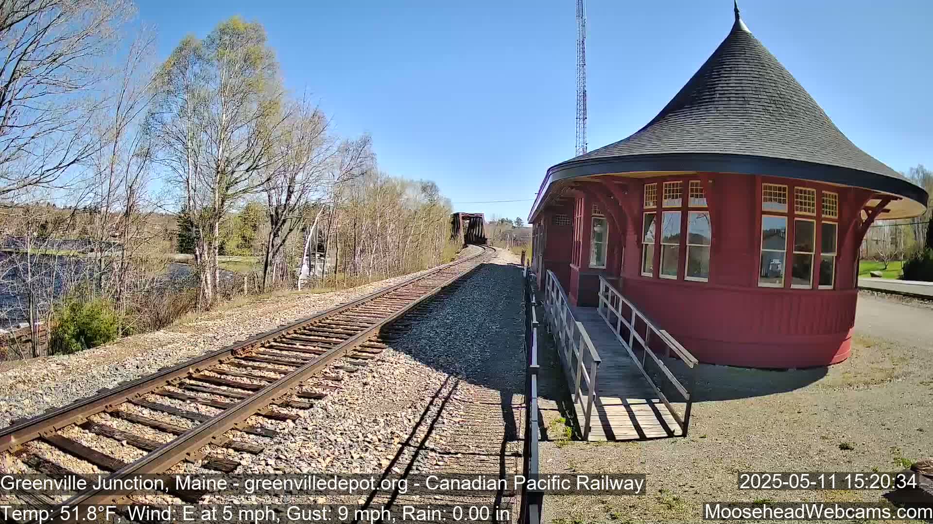 A red, round train depot with a conical roof sits beside railroad tracks that lead to a bridge, all under a clear blue sky.