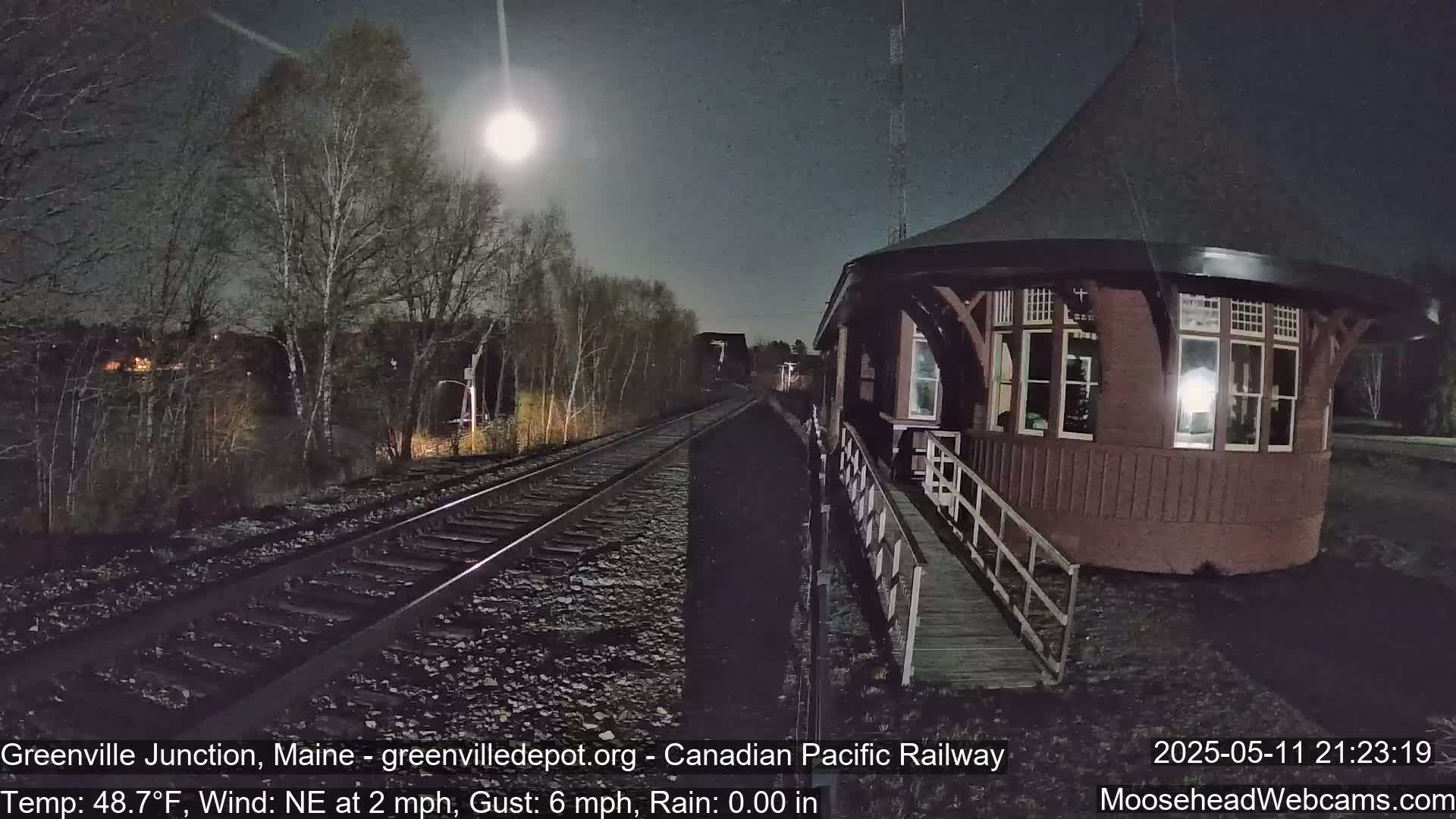 Under a bright moonlit sky, a red octagonal train station stands beside railroad tracks at night.