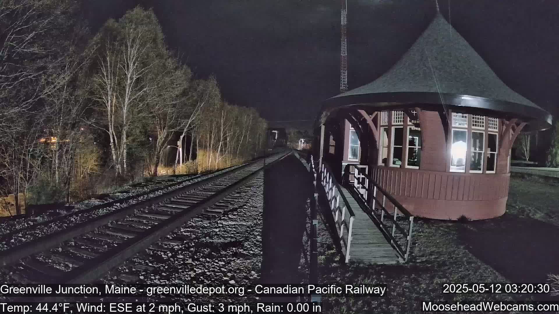 A red, round train depot with a walkway is illuminated at night next to railroad tracks that disappear into a dark, tree-lined landscape under a clear sky.