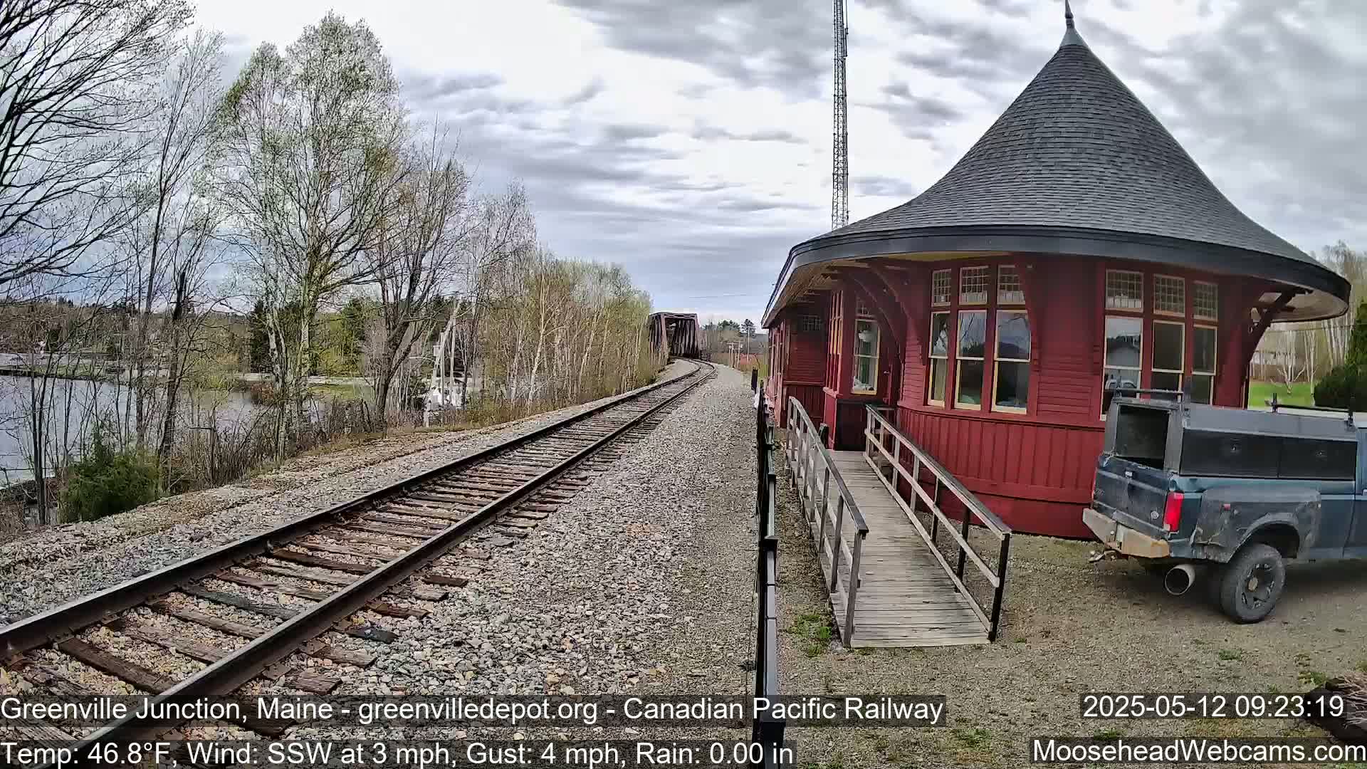 A red train depot with a conical roof sits beside railroad tracks leading to a bridge, under an overcast sky with light rain.