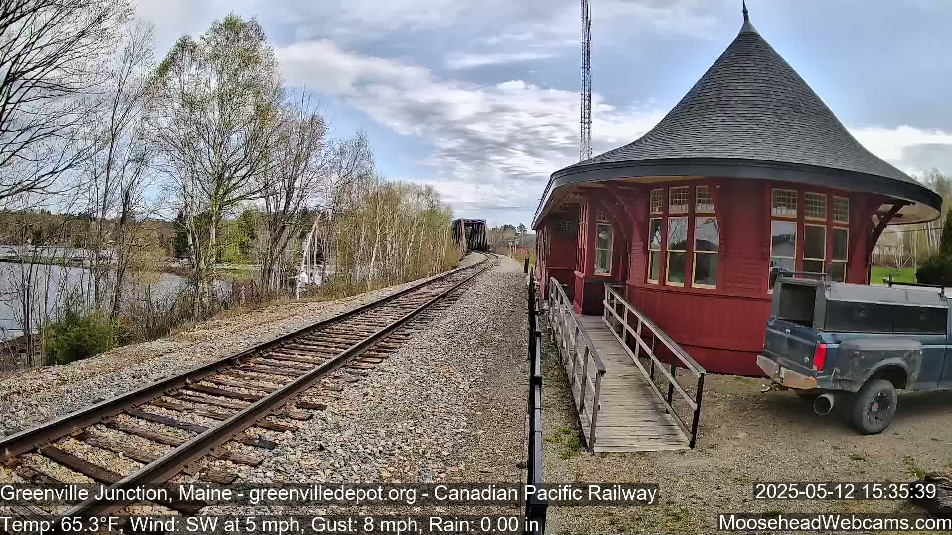 A red, round train station with a ramp sits beside railroad tracks that stretch toward a bridge over a calm body of water under a partly cloudy sky.