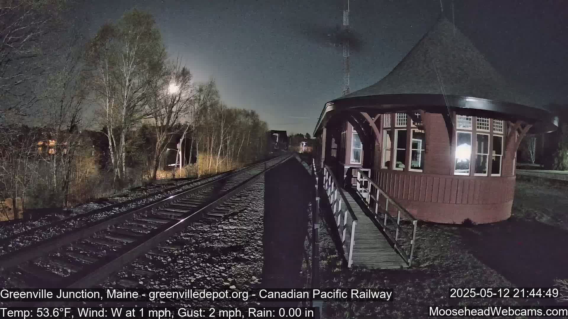 A moonlit night scene shows a red train station beside railroad tracks that extend into a dark, tree-lined landscape under a mostly clear sky, with a light west wind.