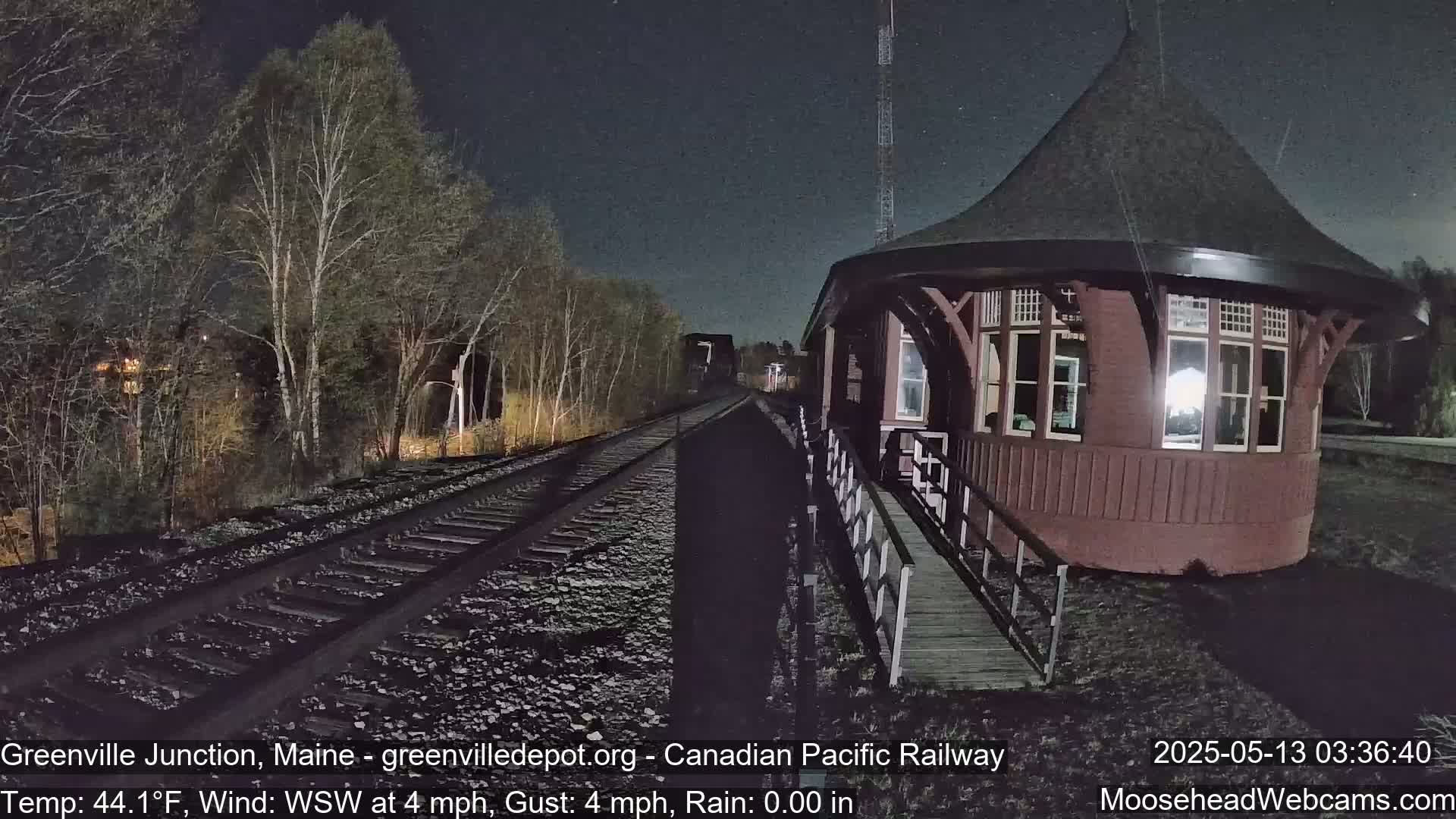 A dark red, round train depot stands beside train tracks under a nighttime sky with light rain and trees in the background.