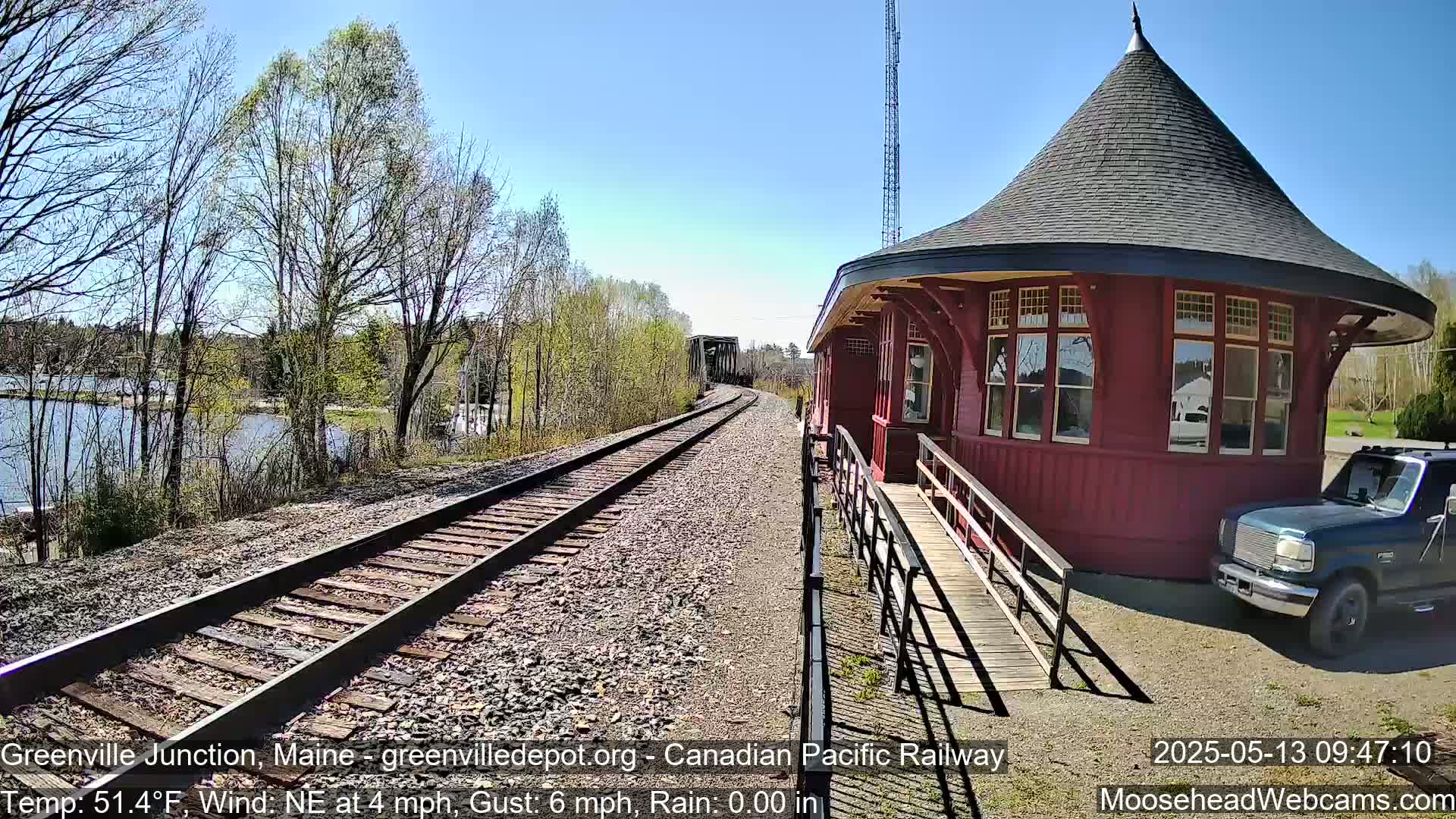 A red, octagonal train station with a conical roof sits beside railroad tracks on a sunny day, with a body of water and trees visible in the background.