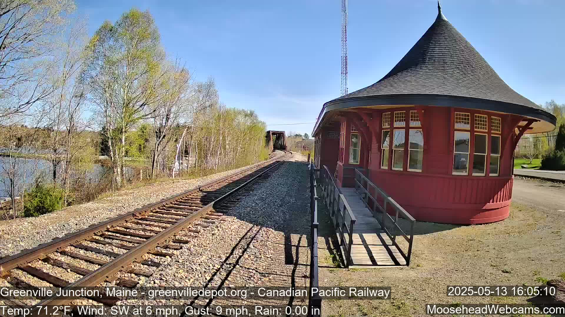 A red, round train depot with a conical roof sits beside railway tracks leading to a bridge, under a sunny sky.