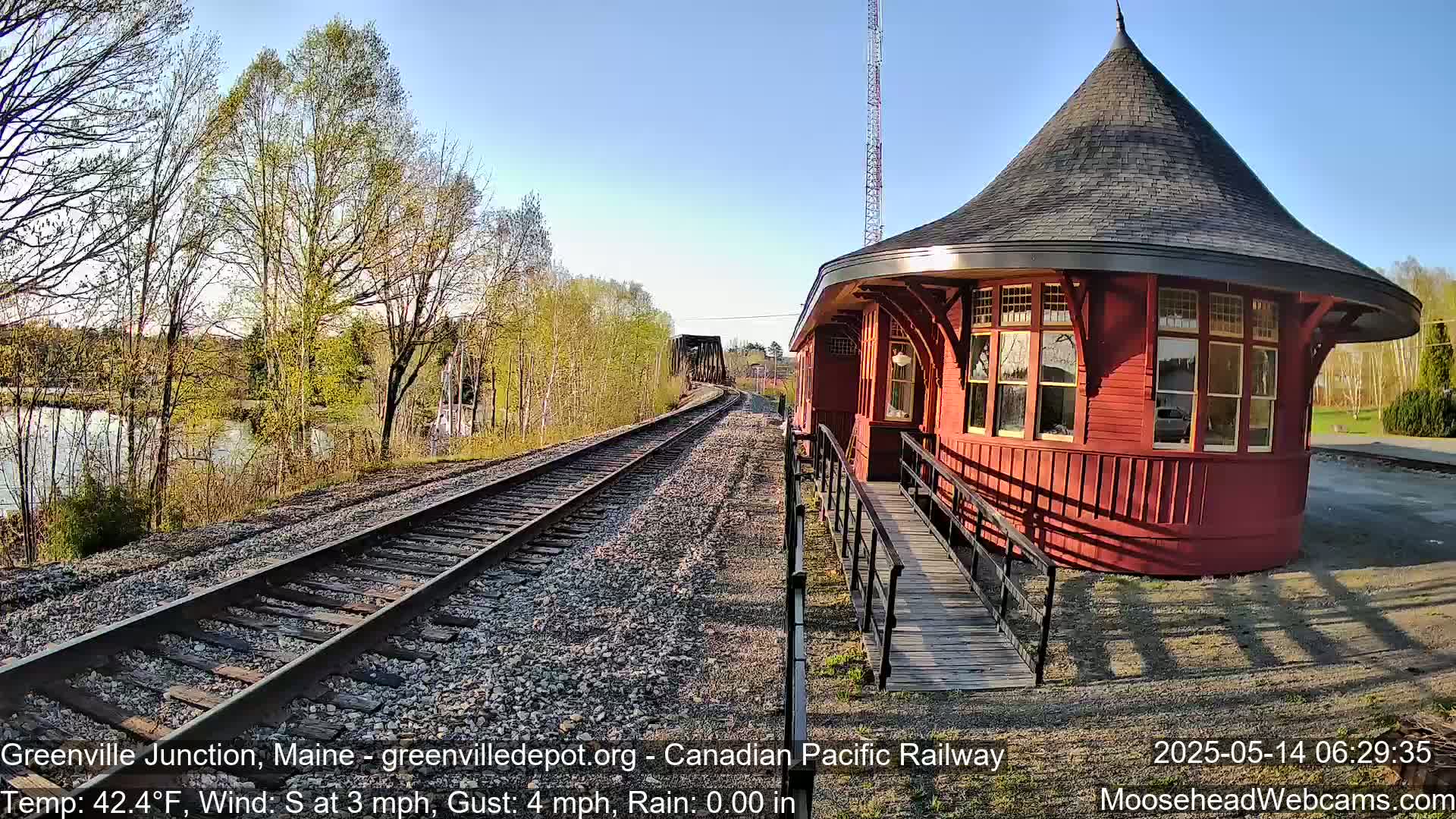 A red, round train station with a conical roof sits beside railroad tracks on a sunny day with a light south wind.