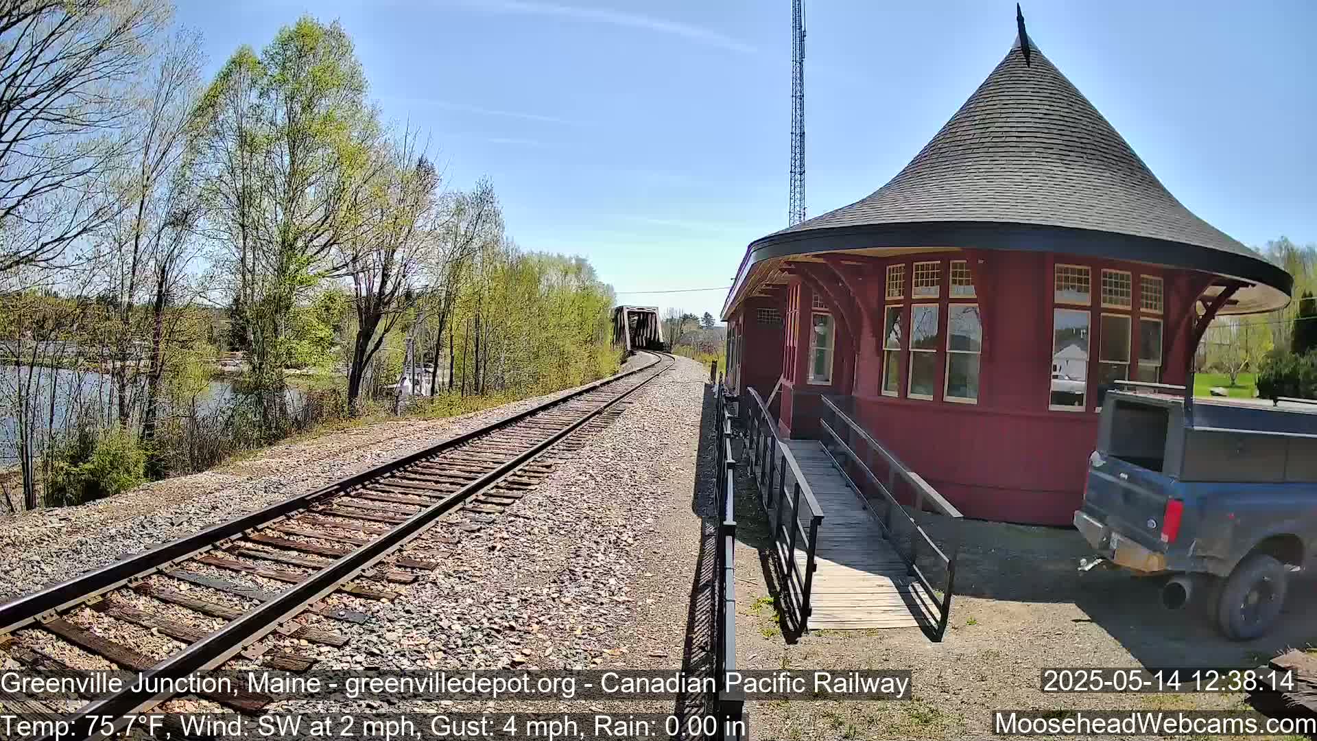 A red, octagonal train station with a conical roof sits beside railroad tracks that lead to a bridge over a calm body of water, under a sunny sky with light winds.