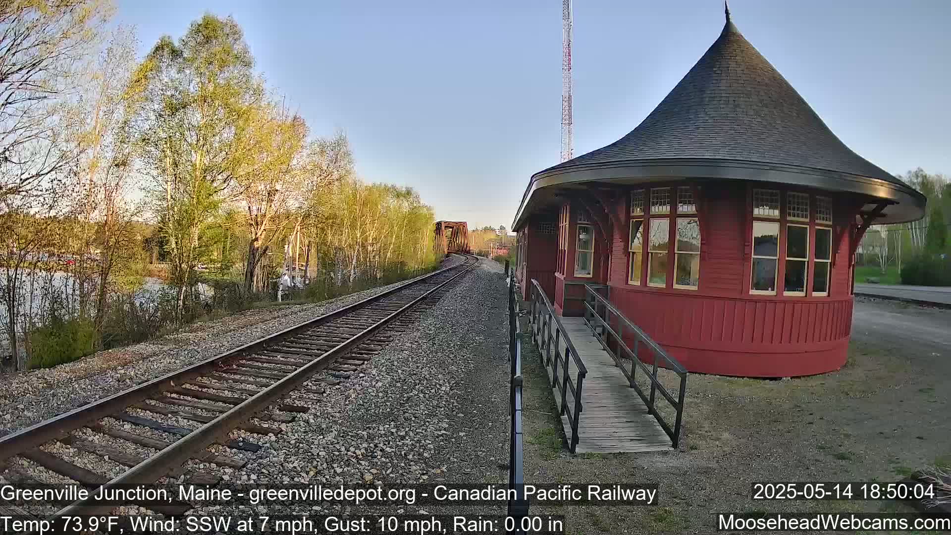 A red, round train depot with a conical roof sits beside railroad tracks that curve away from the camera, passing by a calm body of water and trees under a clear blue sky.