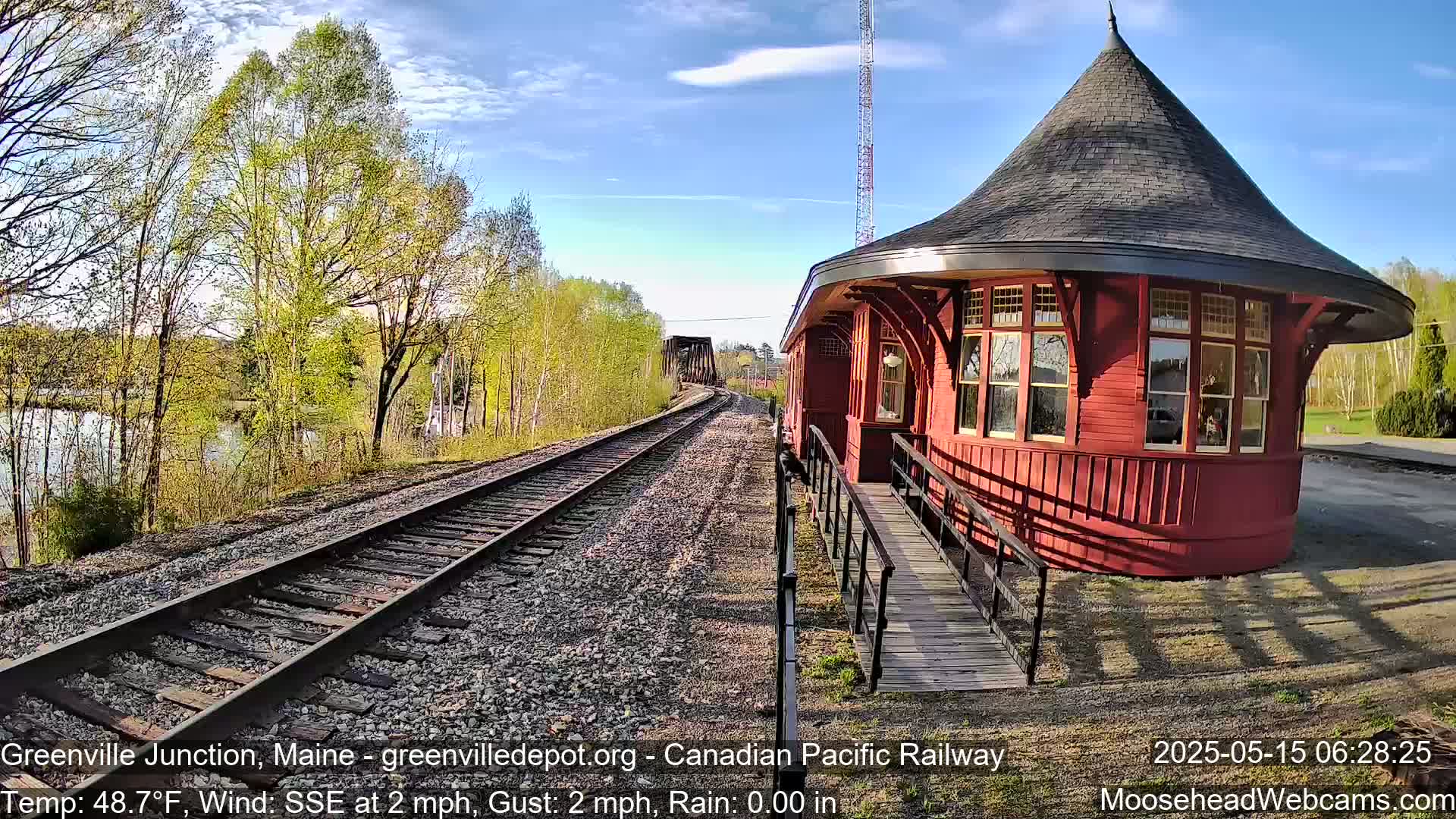 A red, round train depot with a conical roof sits beside train tracks that lead to a bridge over a calm river under a partly cloudy sky.