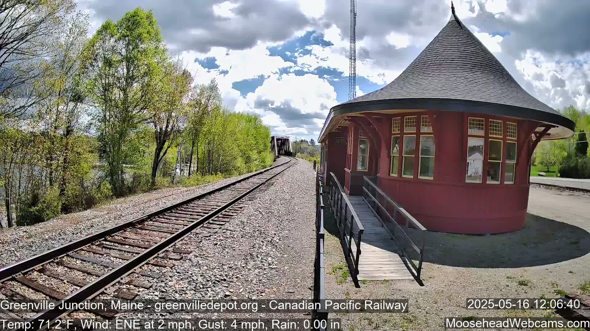 A red, round train depot with a conical roof sits beside railroad tracks leading to a bridge under a partly cloudy sky.