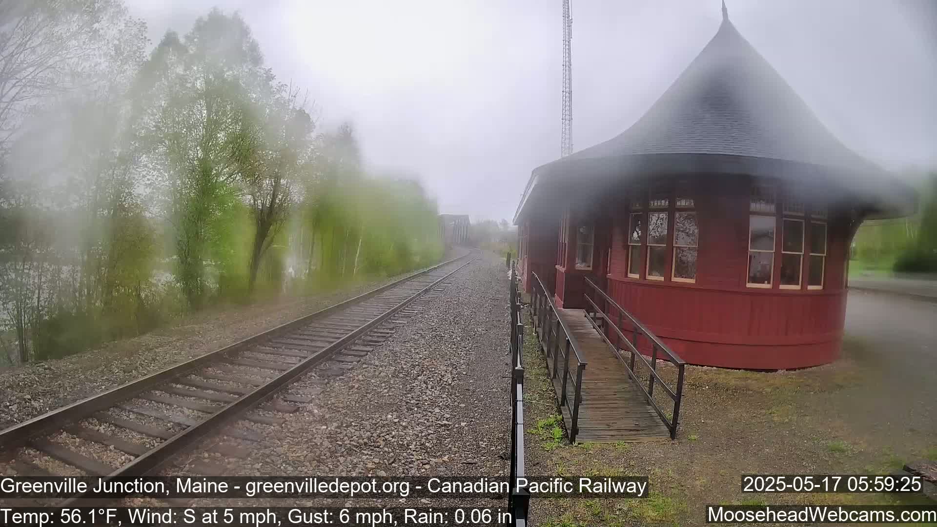 A red, round train station stands beside railroad tracks on a rainy day, with trees and mist visible in the background.