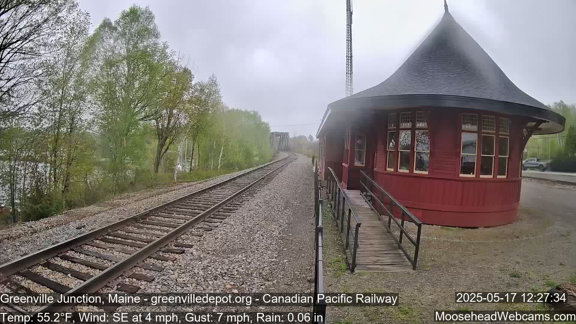 A red, round train station with a conical roof sits beside train tracks on a rainy day, with trees and a river visible in the background.
