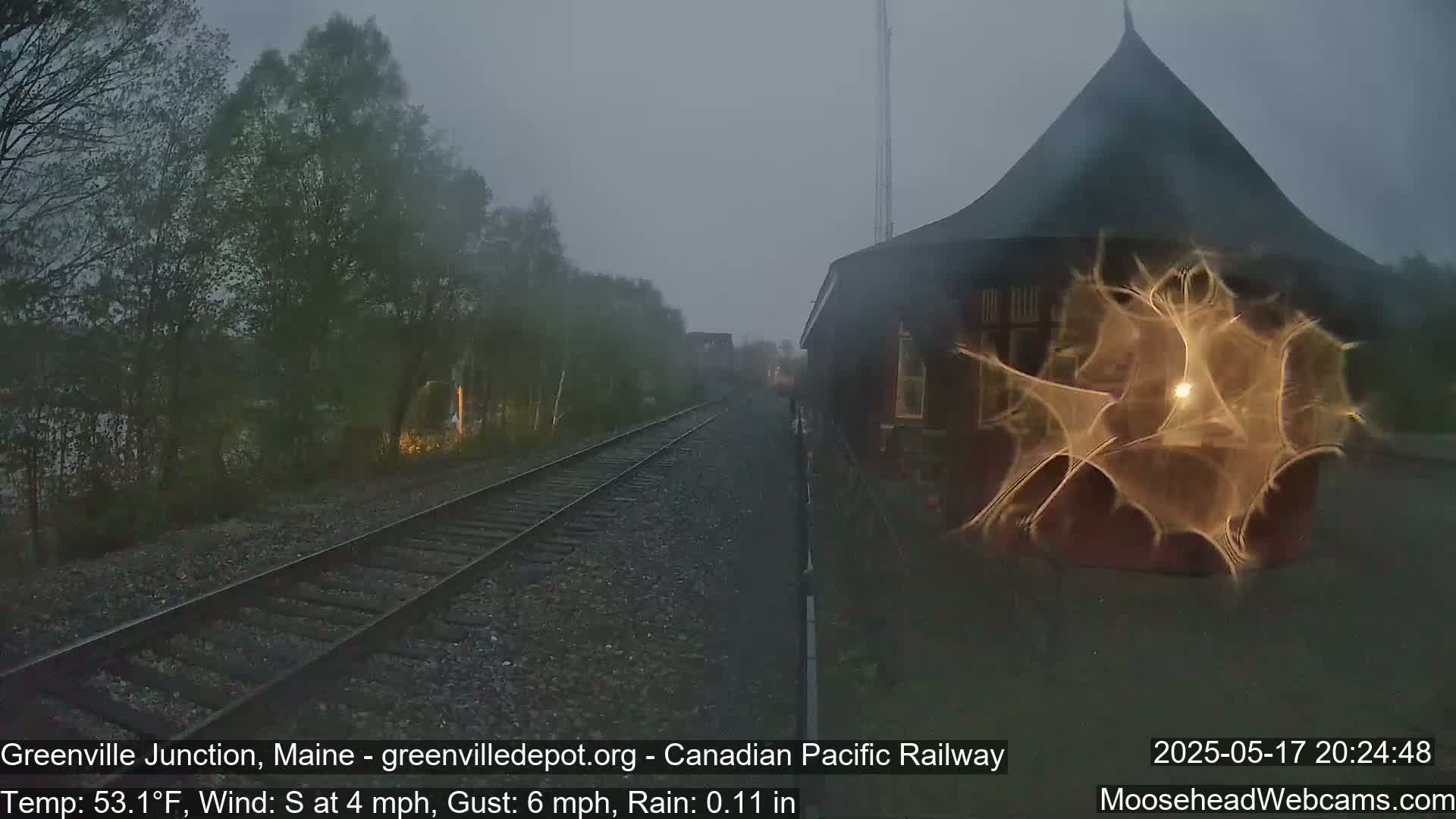 A rain-swept view of railroad tracks extending toward a dark, conical-roofed building under a gloomy sky.