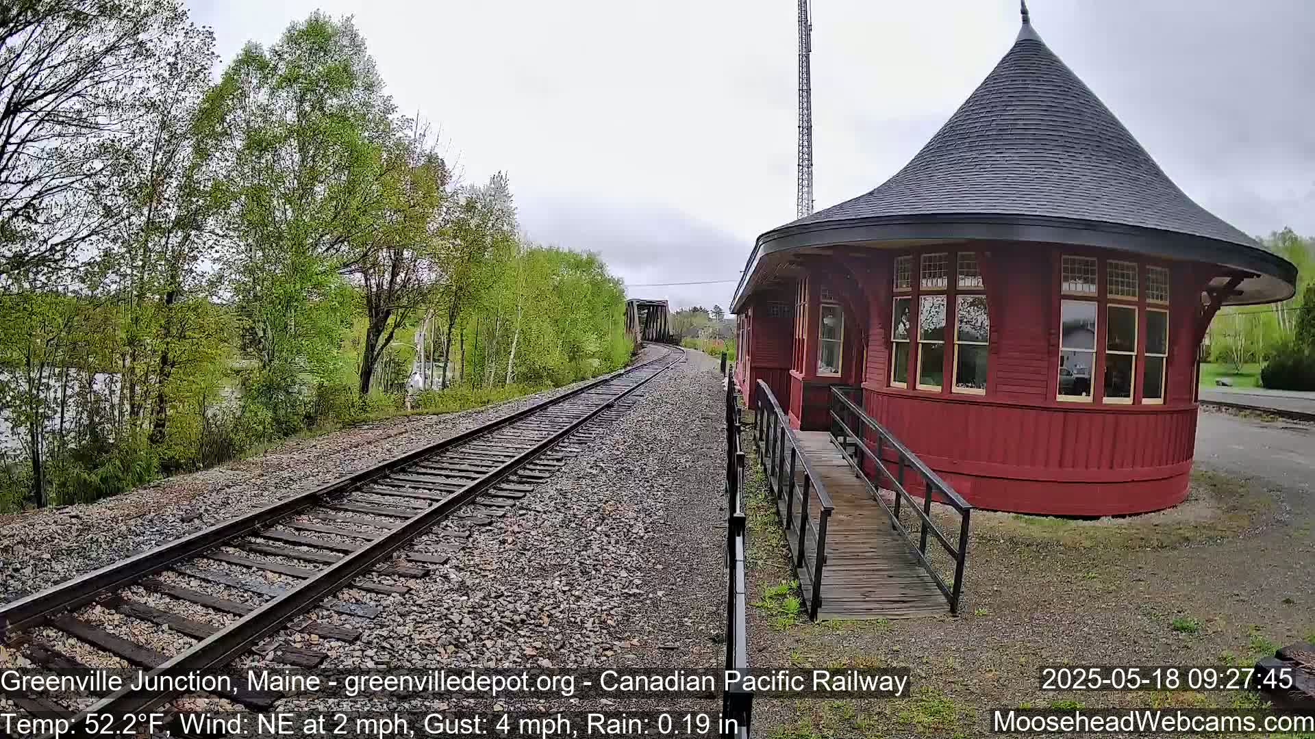 A red, octagonal train depot with a conical roof sits beside railroad tracks curving into the distance under an overcast sky, with light rain falling and lush green trees nearby.