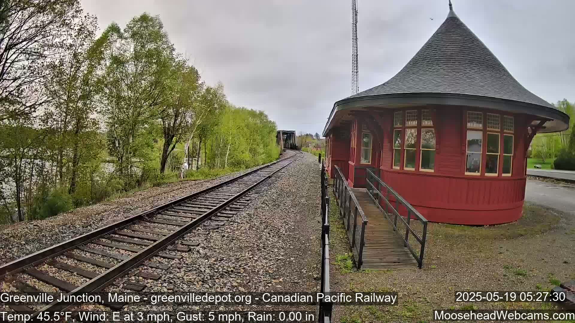 A red, round train depot with a conical roof stands beside railroad tracks that lead to a distant bridge, under an overcast sky with a slight east wind.