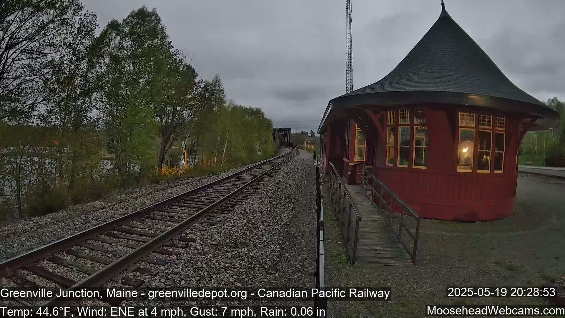 A red, round train station with a conical roof is situated beside railway tracks leading to a bridge, under a cloudy, rainy sky.