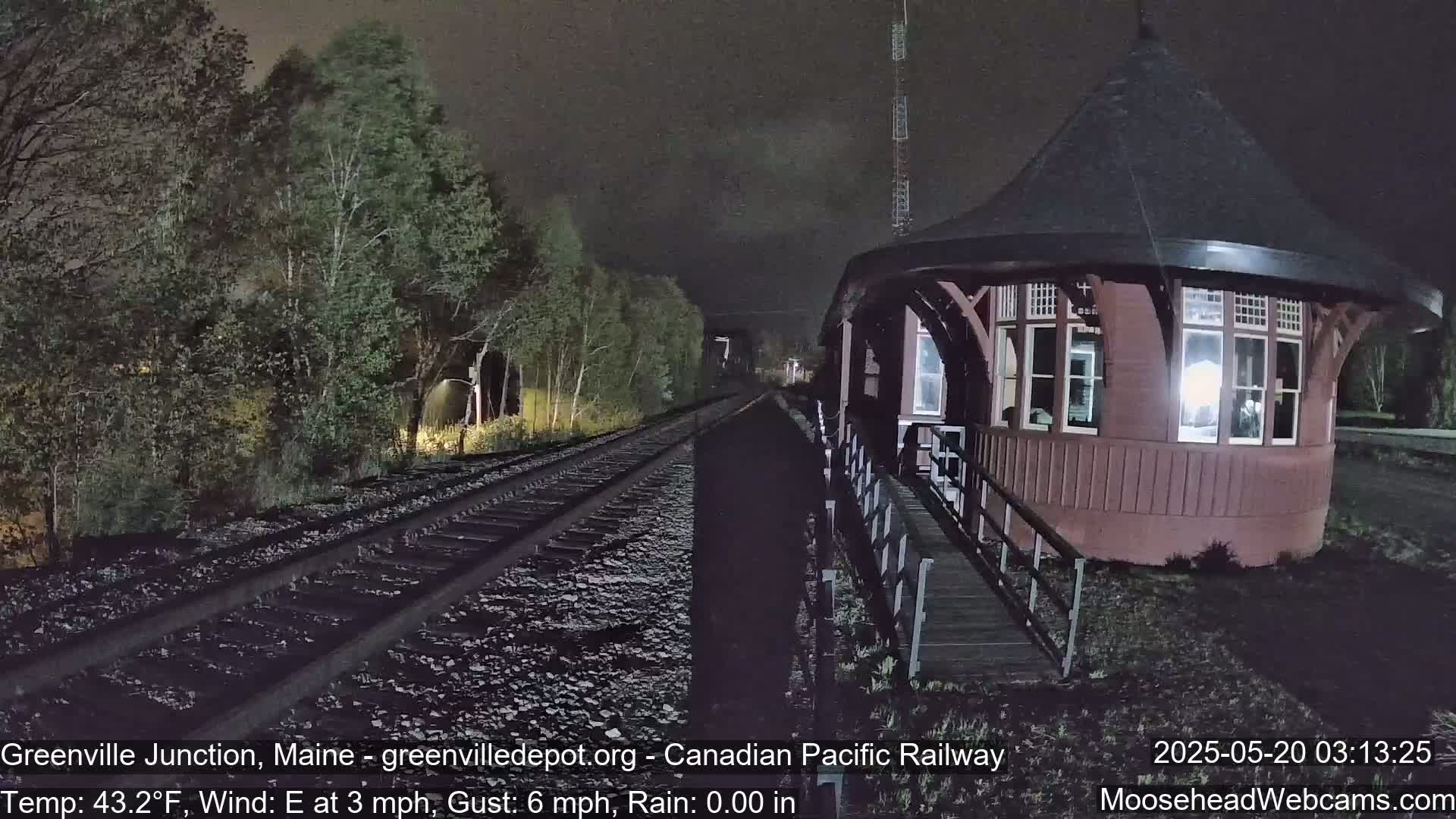 A dark red, octagonal train station sits beside railroad tracks at night, under a mostly clear sky, with trees lining the tracks.