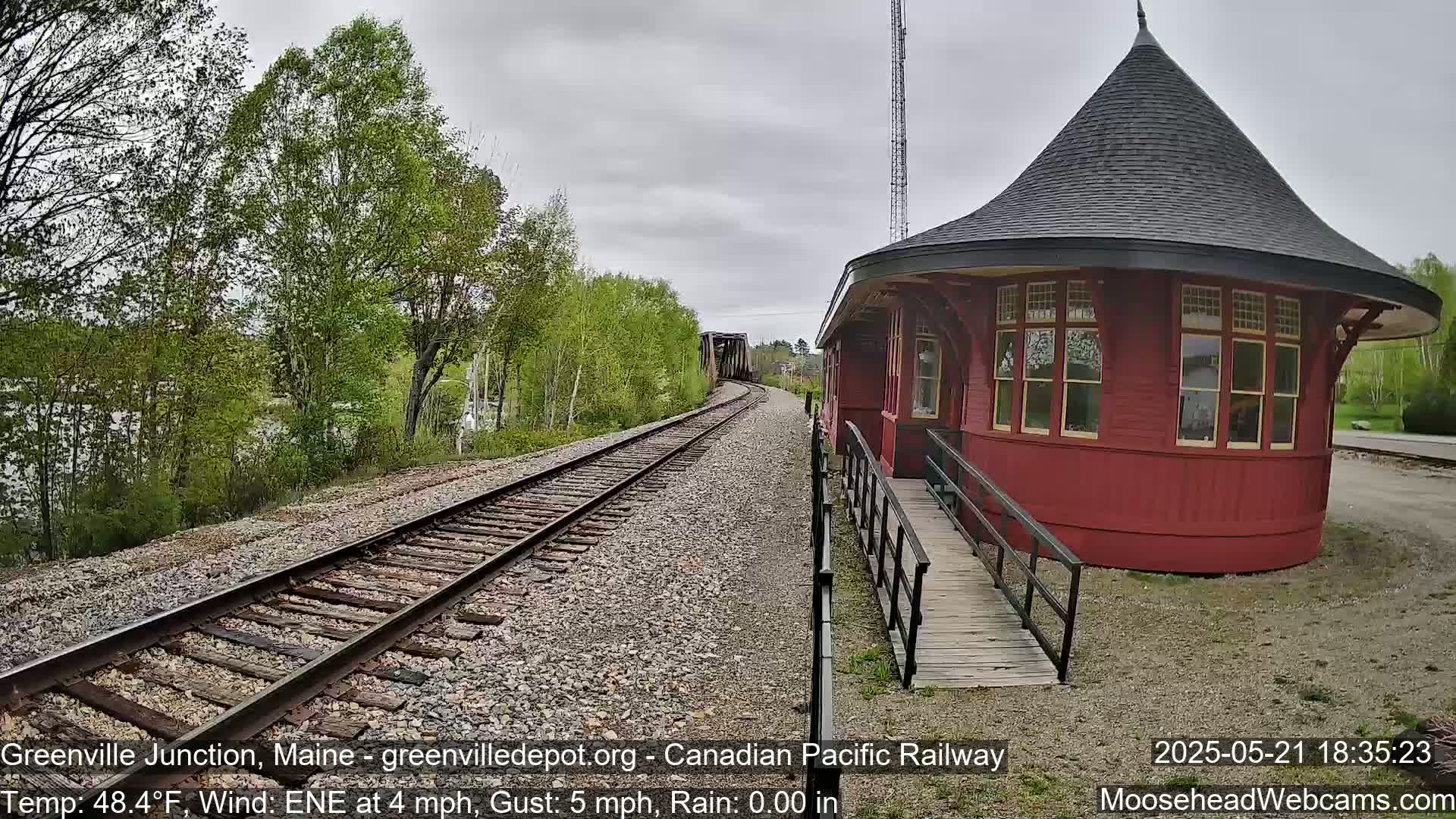 A red octagonal train depot with a conical roof sits beside train tracks that curve toward a bridge under a cloudy sky.