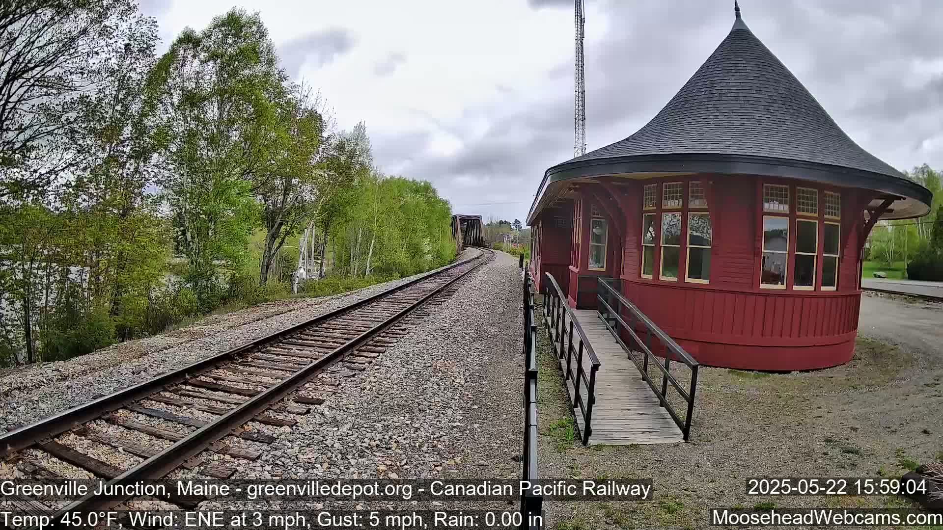 A red, round train depot with a conical roof sits beside railroad tracks curving toward a bridge under a cloudy sky.