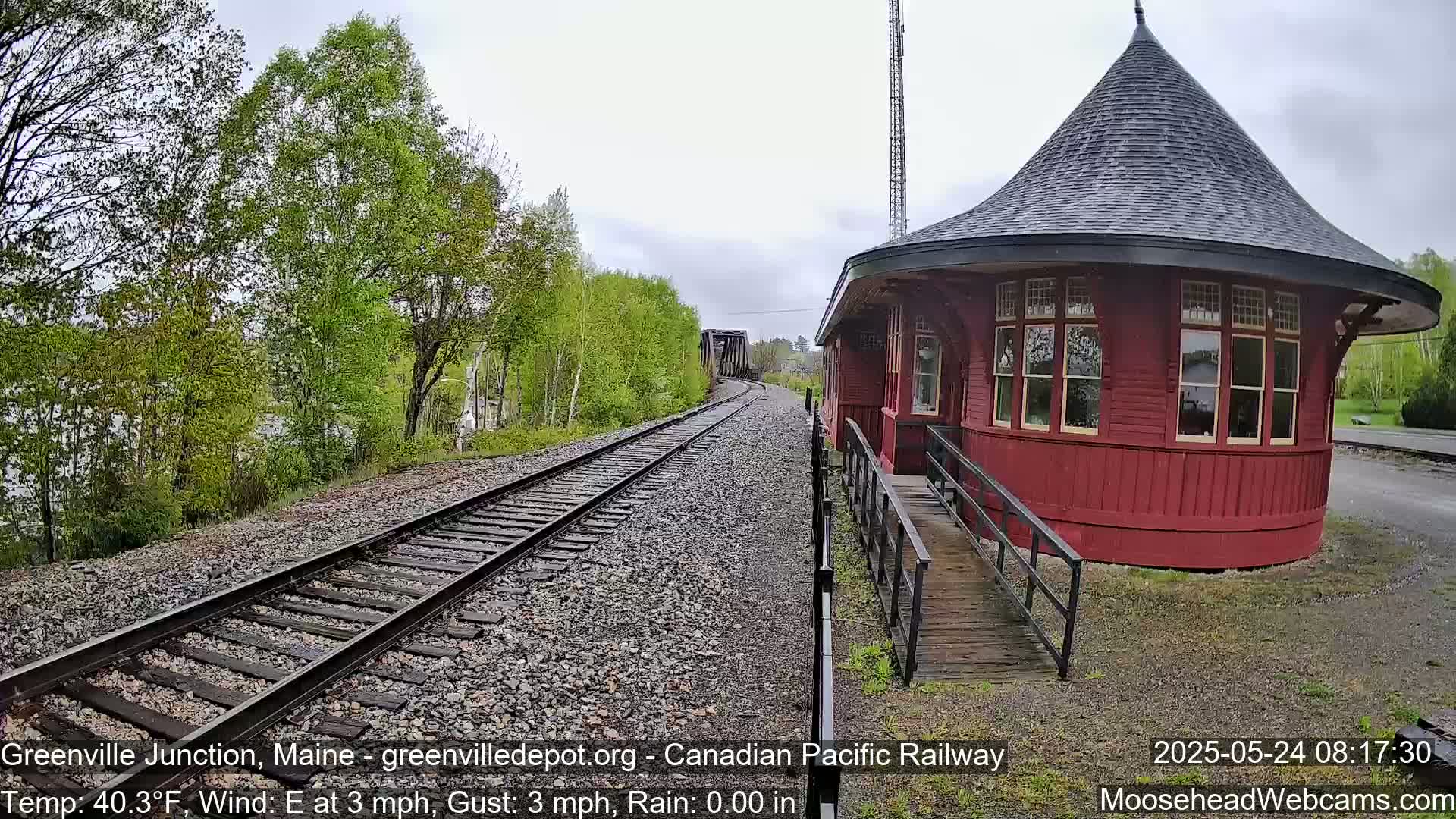Train tracks extend toward a red, round train depot next to a body of water under a cloudy sky.