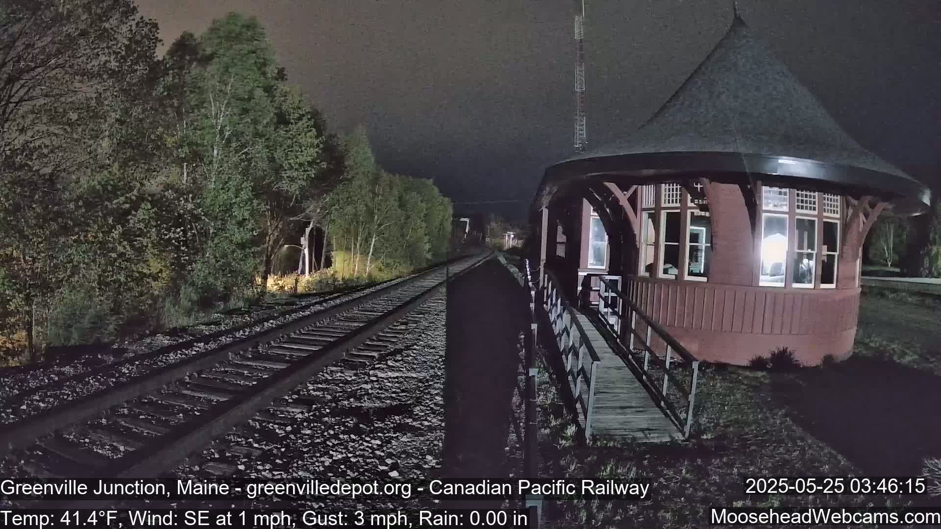 Train tracks stretch into the night, flanked by trees, leading to a dark red, round train depot under a dark, slightly cloudy sky.