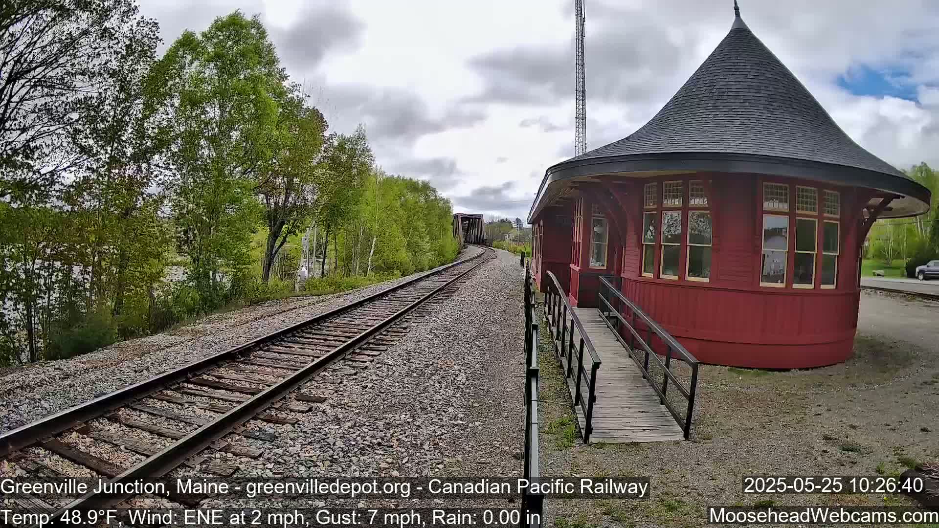 Train tracks extend toward a red, round train depot under a cloudy sky, with green trees lining the tracks.