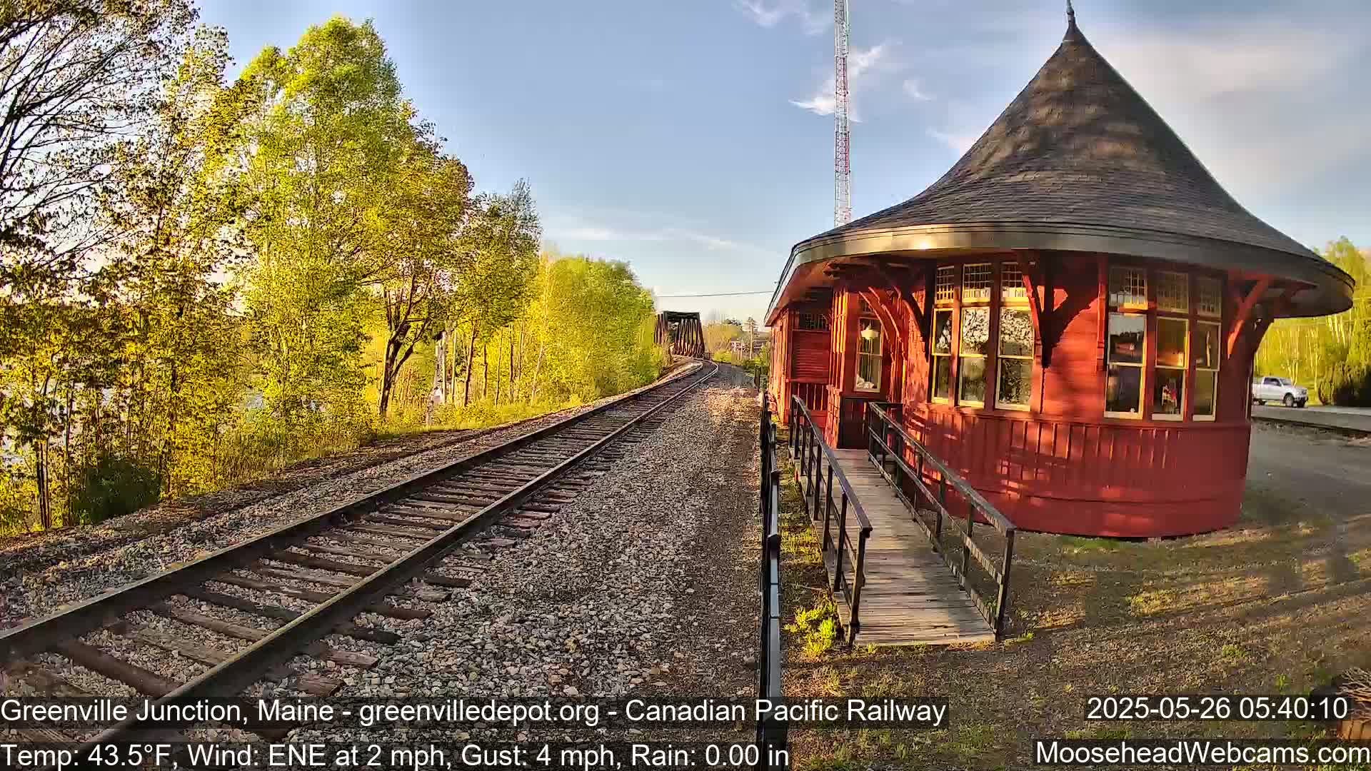 A red, round train station with a ramp sits beside railroad tracks that curve toward a bridge under a partly sunny sky, with light green trees lining the tracks.