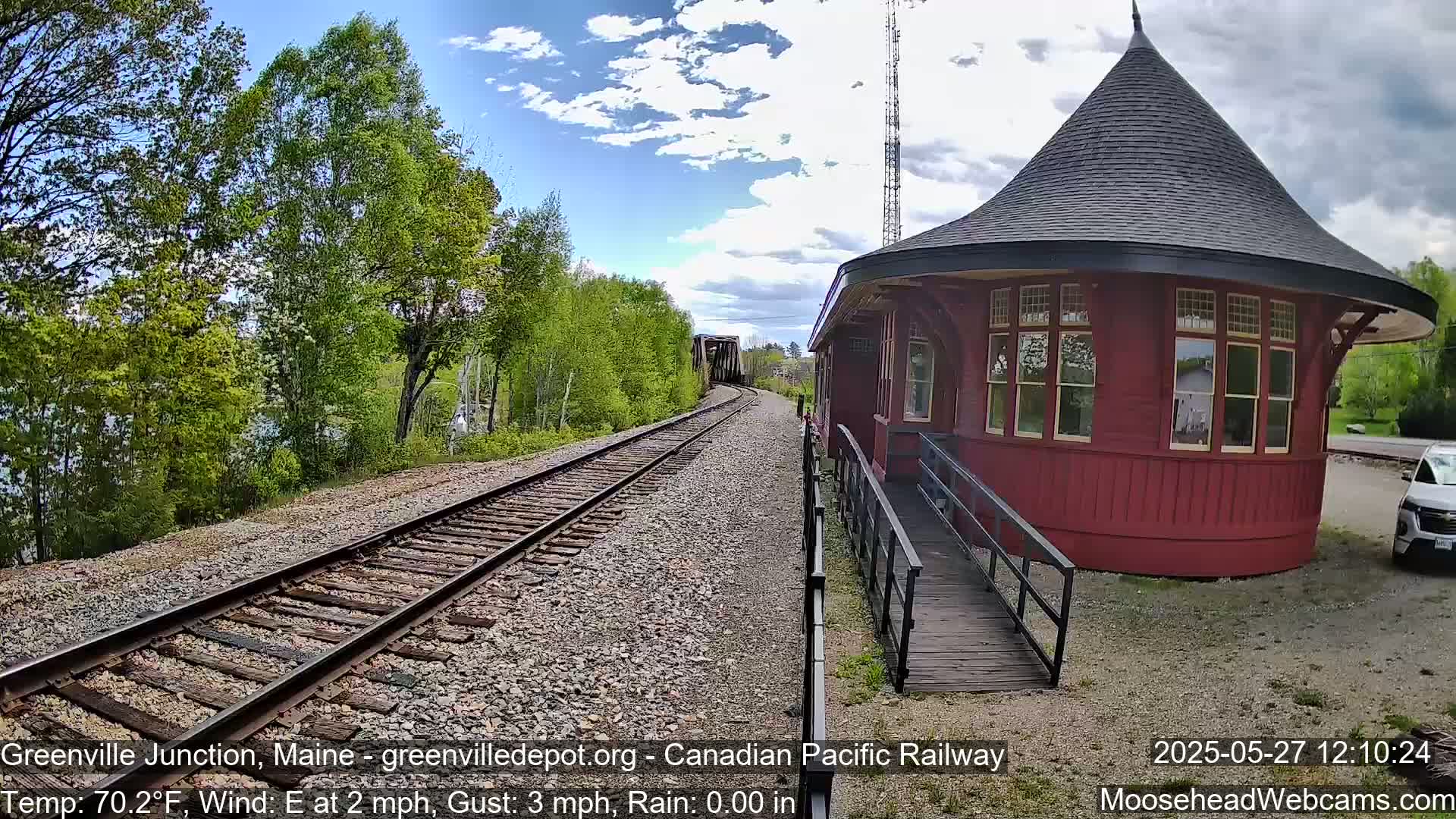 A red, round train station with a ramp sits beside railroad tracks that lead toward a bridge and lush green trees under a partly cloudy sky.