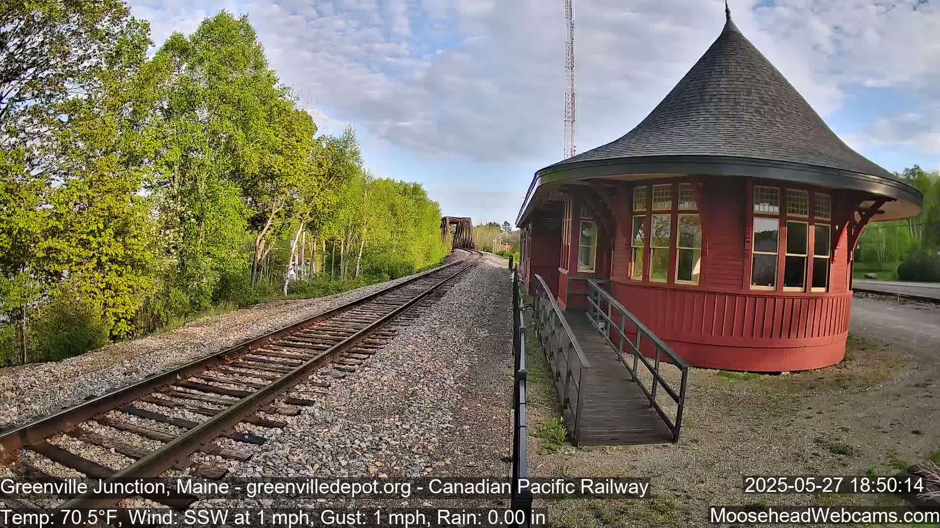 A red, round train depot with a conical roof sits beside train tracks that extend to a bridge in the distance under a partly cloudy sky.