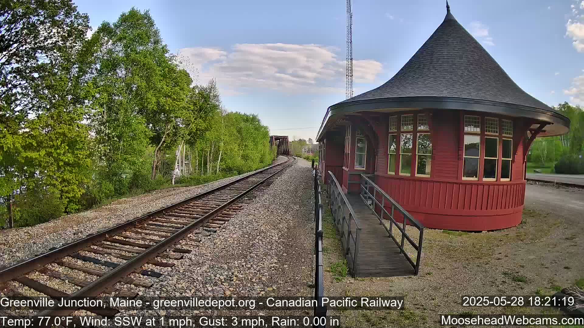 A red, round train depot with a conical roof sits beside railroad tracks that curve towards a distant bridge under a partly cloudy sky, with a light rain.