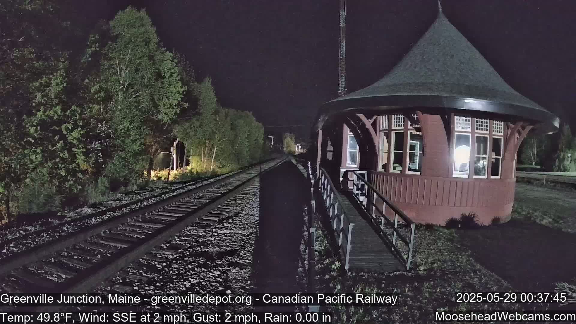 A red, round train station is illuminated at night next to a railway track that runs through a wooded area under rainy conditions.