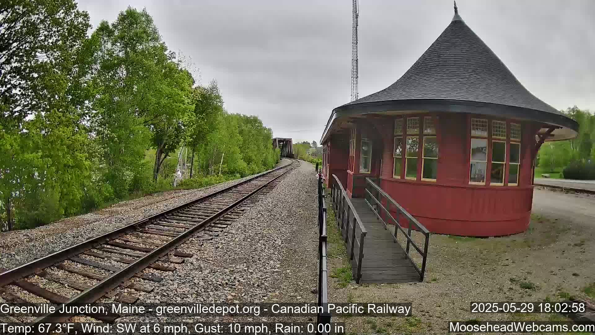 A red, round train station with a conical roof stands beside railroad tracks that curve toward a bridge under an overcast sky.