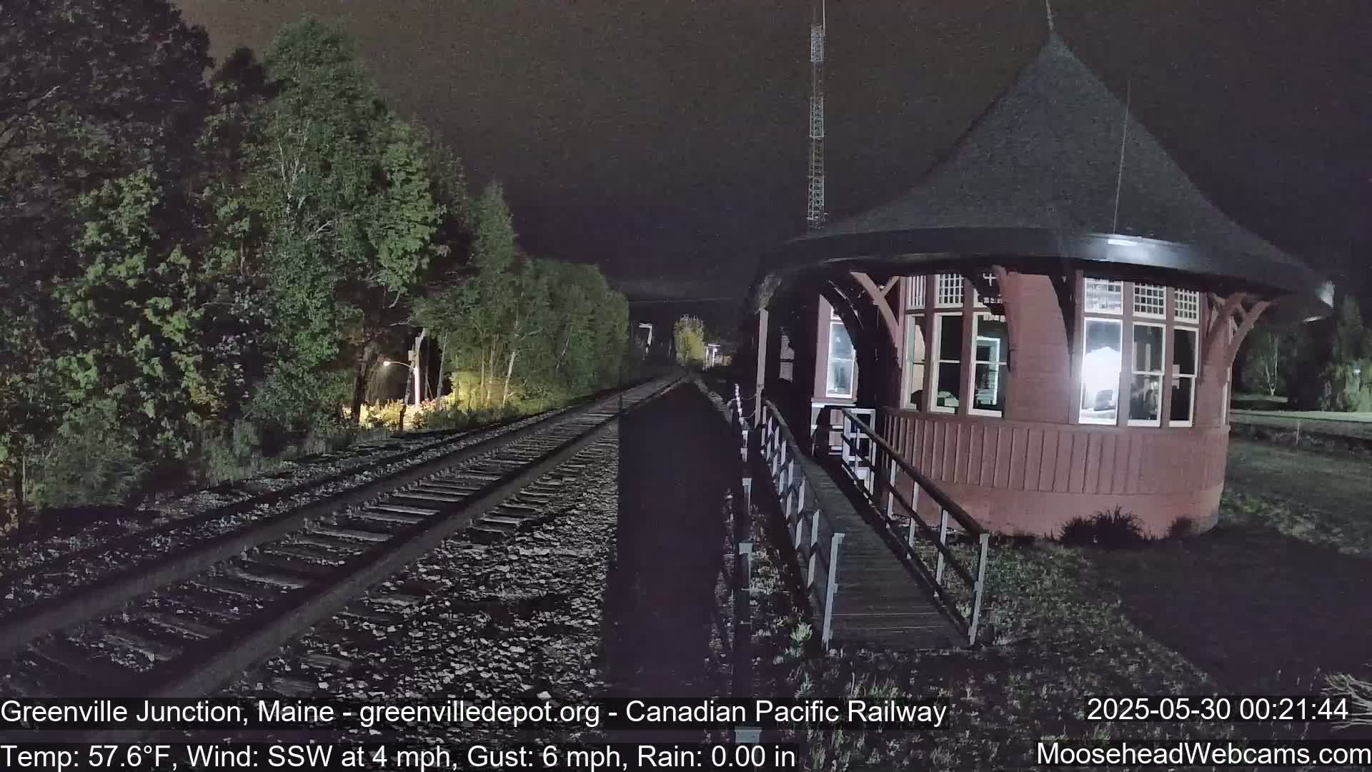 A red, octagonal train depot is shown at night in the rain, next to railroad tracks bordered by trees.