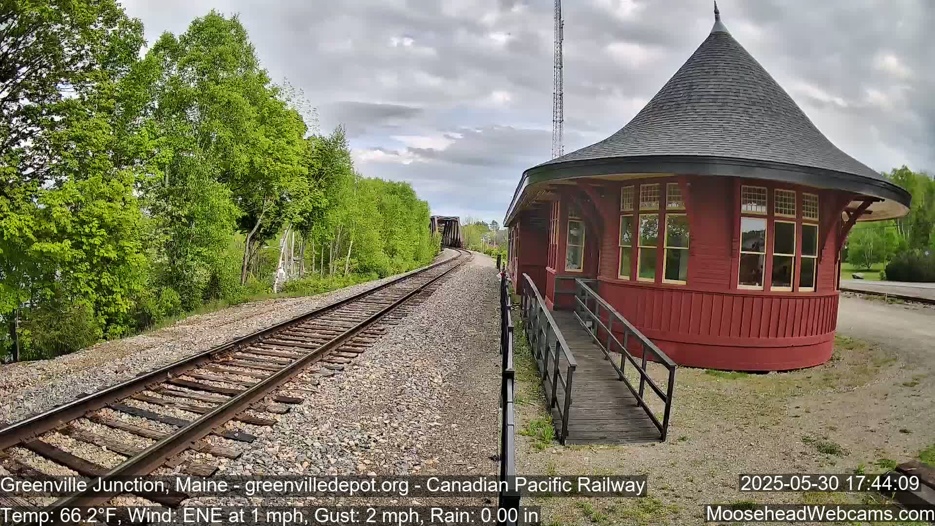 A red, round train depot with a peaked roof sits beside railroad tracks that curve gently to the left, under a partly cloudy sky.