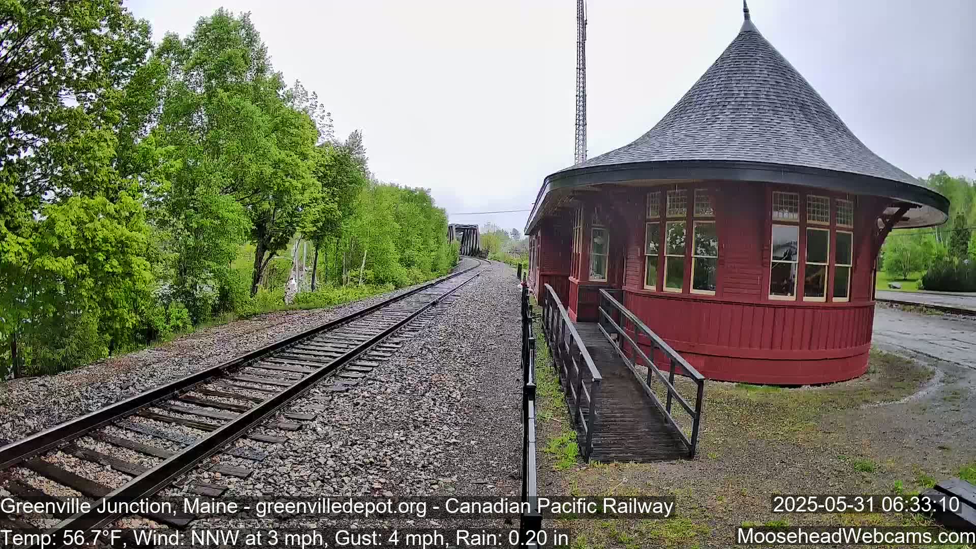 A red, round train depot with a dark roof sits beside train tracks in the rain, surrounded by green trees.
