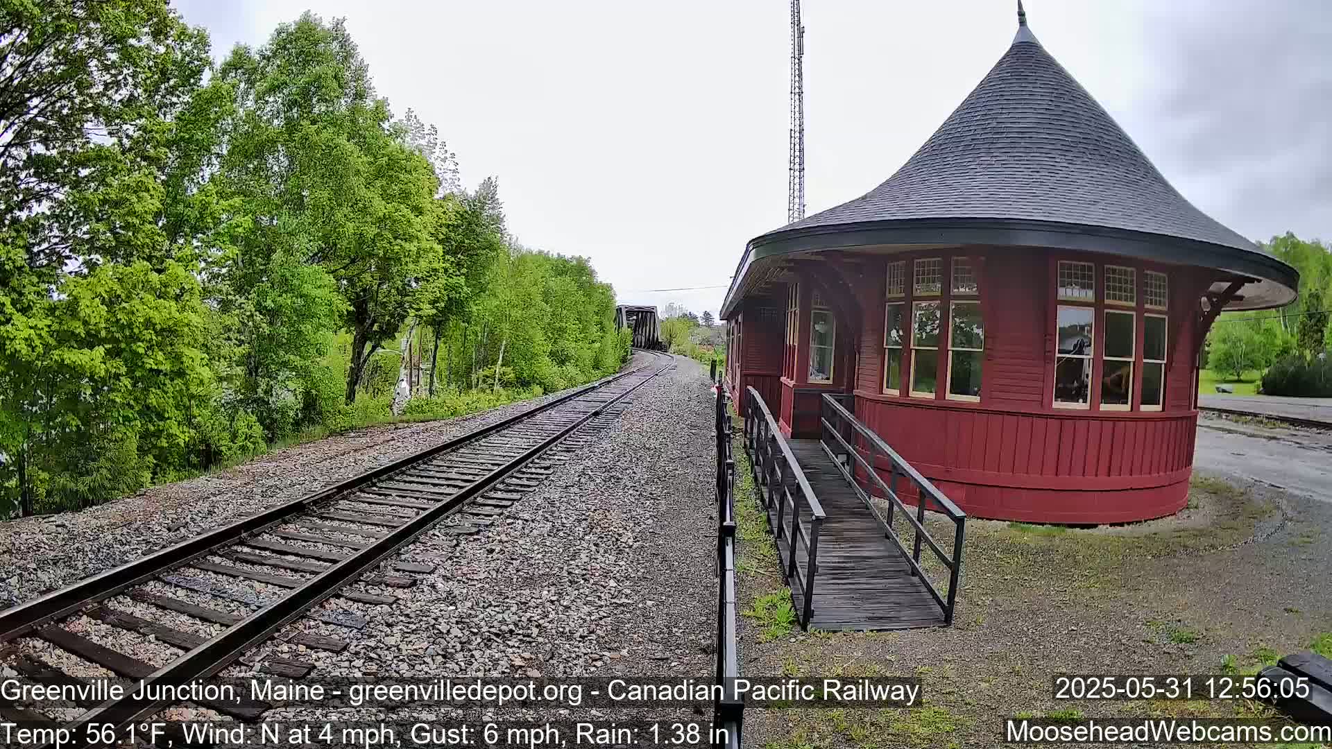 A red, round train station with a ramp is situated beside railway tracks that curve into a wooded area under rainy conditions.