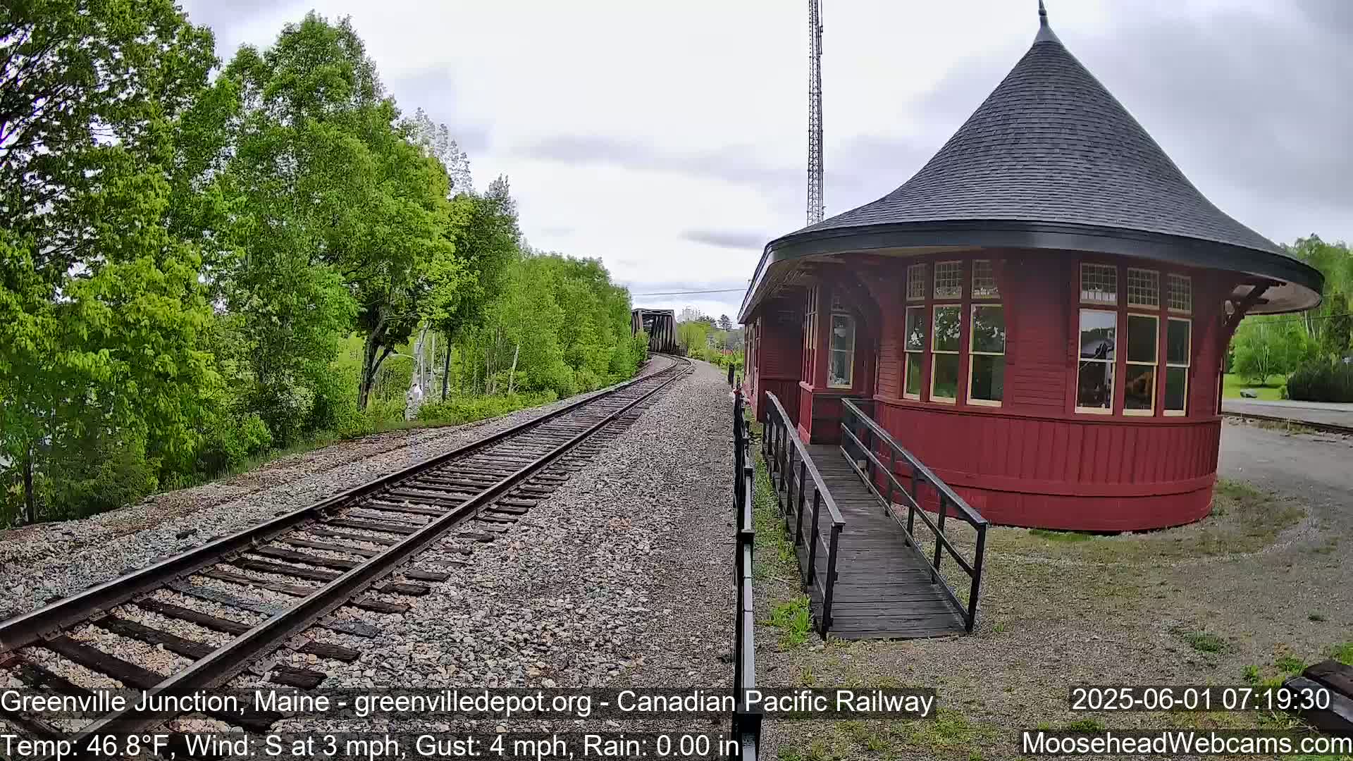A red, octagonal train station with a conical roof sits beside railroad tracks extending into a distance where a train is partially visible under a partly cloudy sky.