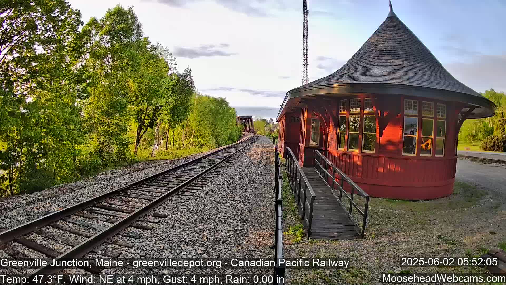 A red, round train depot with a conical roof sits beside railroad tracks on a partly sunny day with a gentle northeast wind.