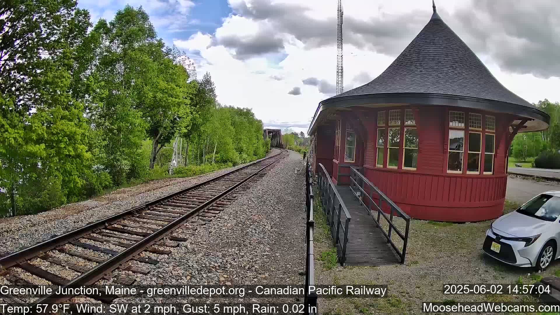 A red octagonal train depot with a conical roof is situated next to train tracks on a slightly rainy day with a light southwest wind.
