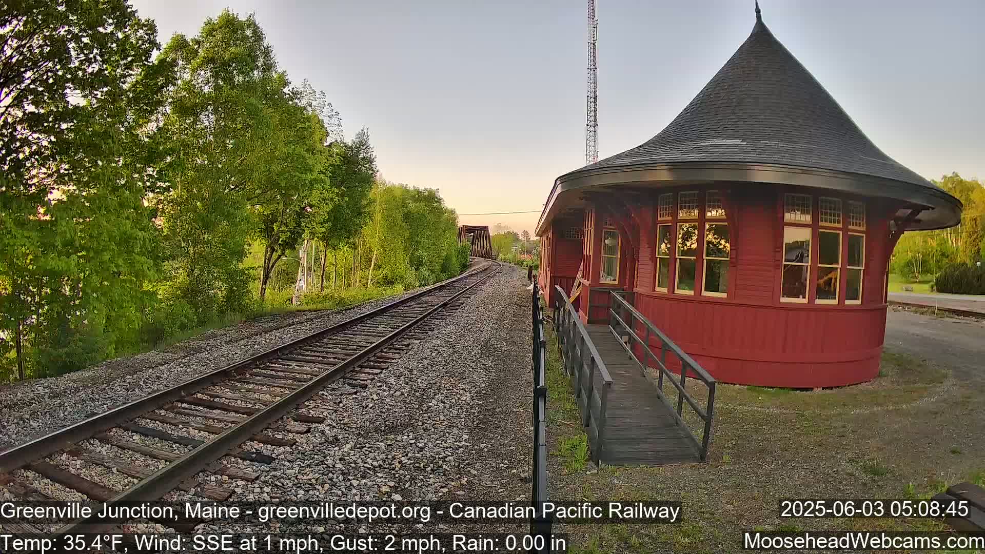 A red, round train station with a conical roof sits beside train tracks that disappear into the distance under a clear, twilight sky, next to a line of green trees.