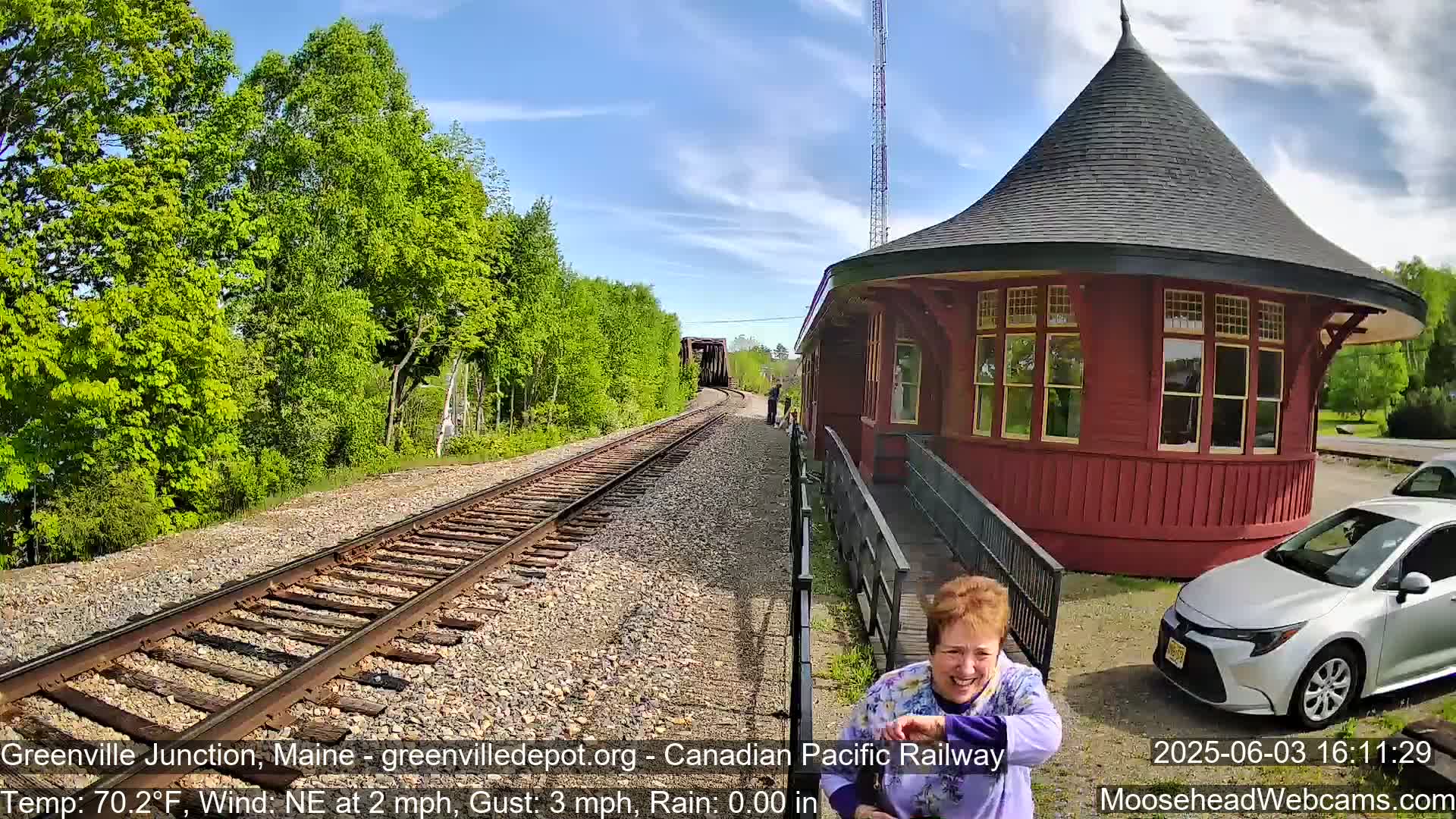 A woman laughs near a red, round train depot on a sunny day with a light northeast wind, next to railroad tracks extending towards a bridge and lush green trees.