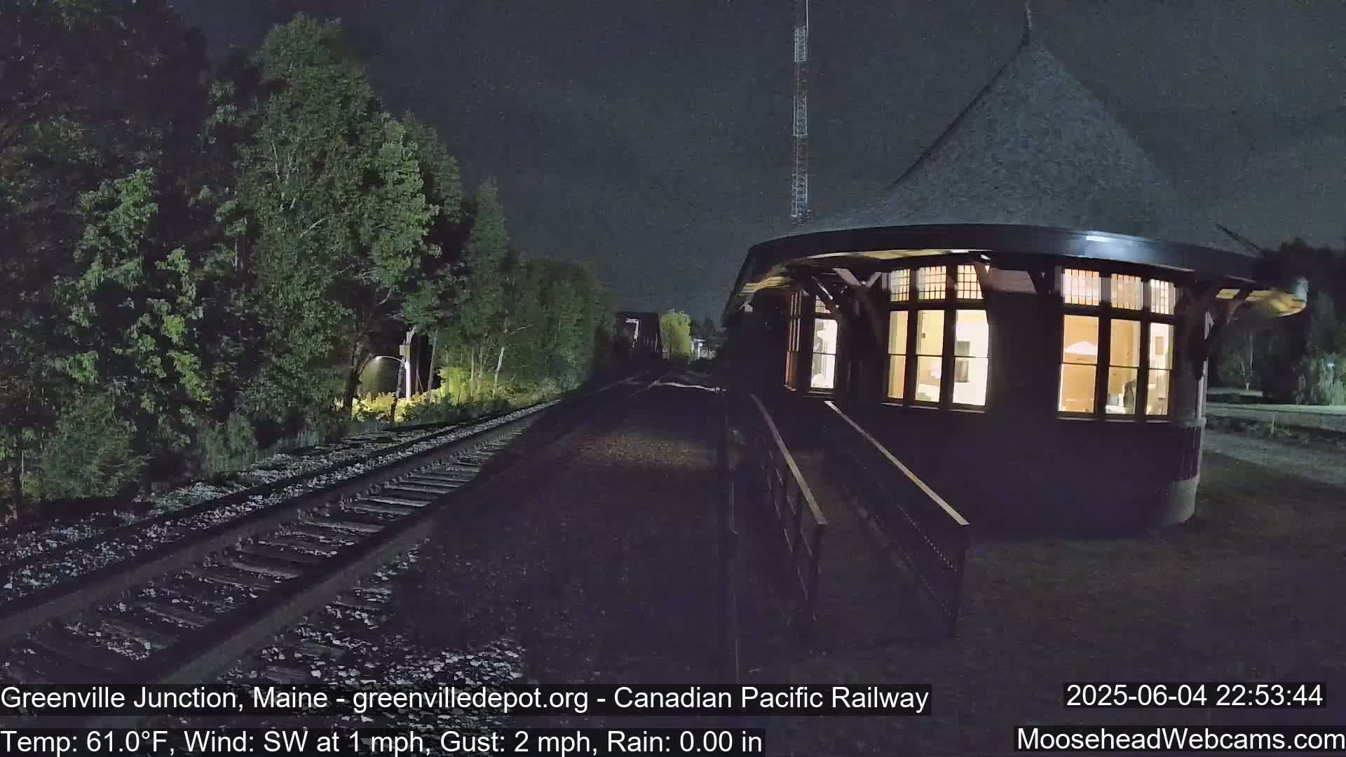 A lit-up, circular train station sits beside railway tracks at night, surrounded by dark green trees under a clear sky.