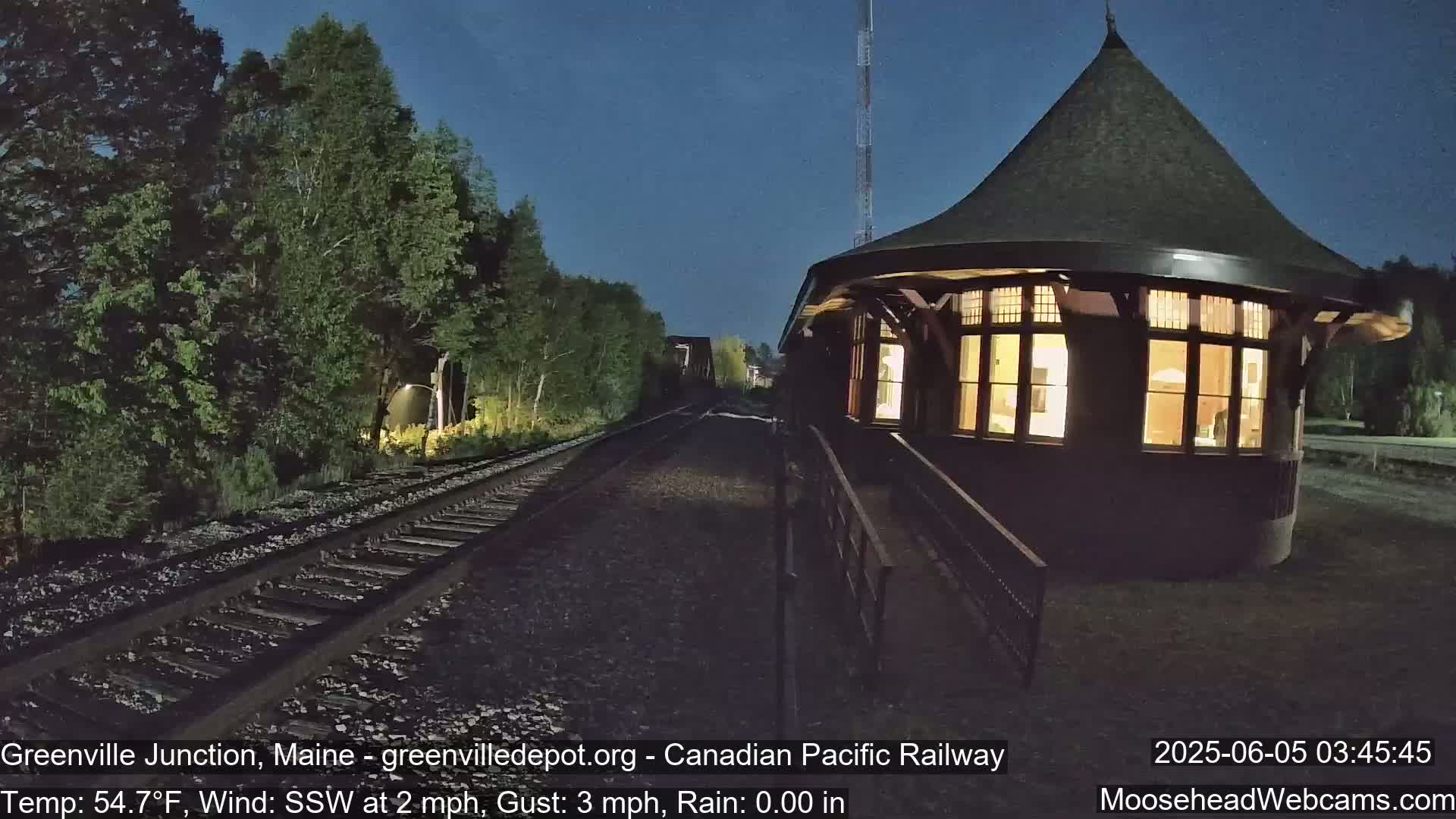 A brightly lit, round train depot stands at night next to train tracks that disappear into a dark, tree-lined area under a clear, dark sky.