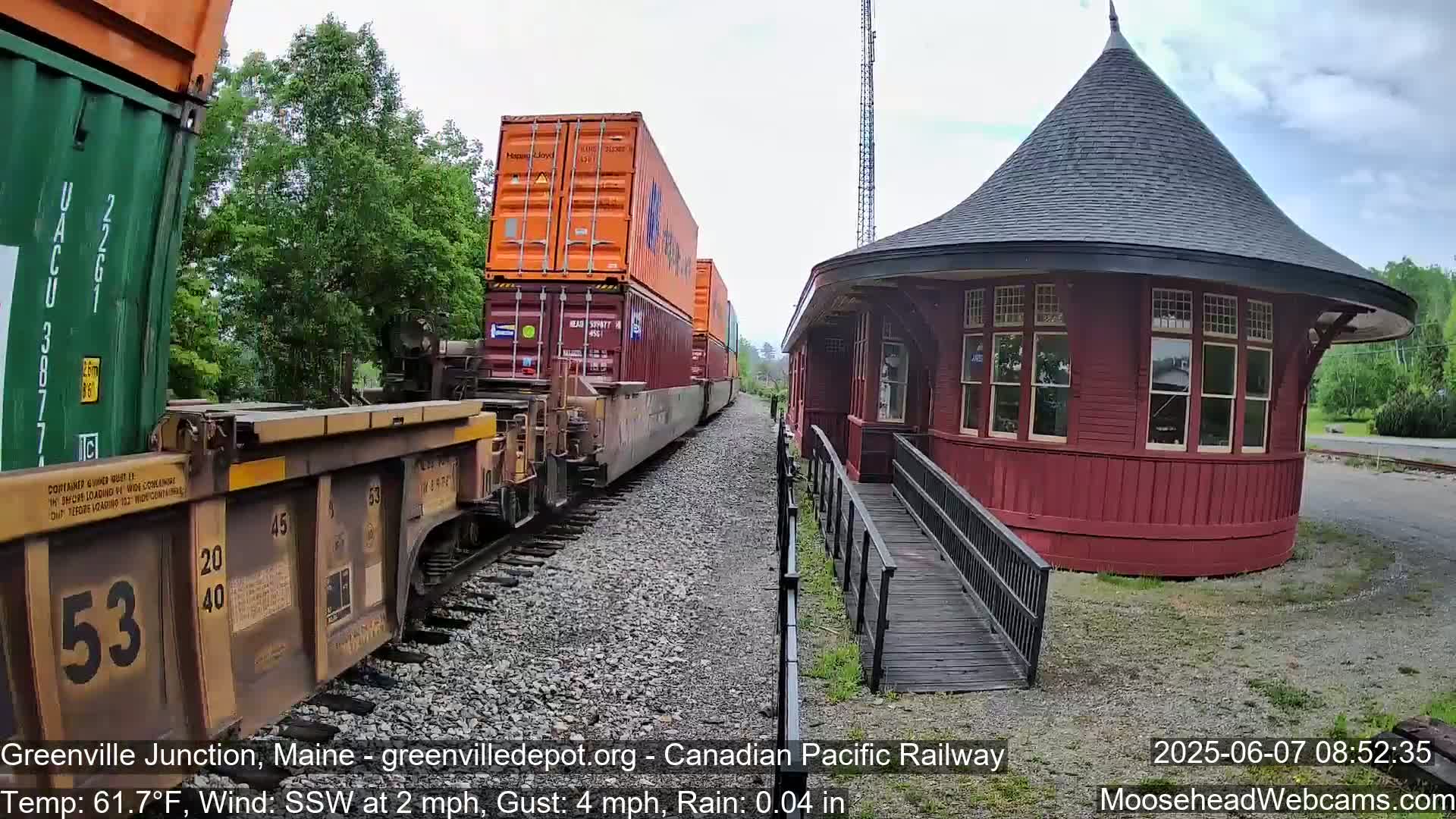 A freight train carrying stacked shipping containers passes a red, round train station in light rain.