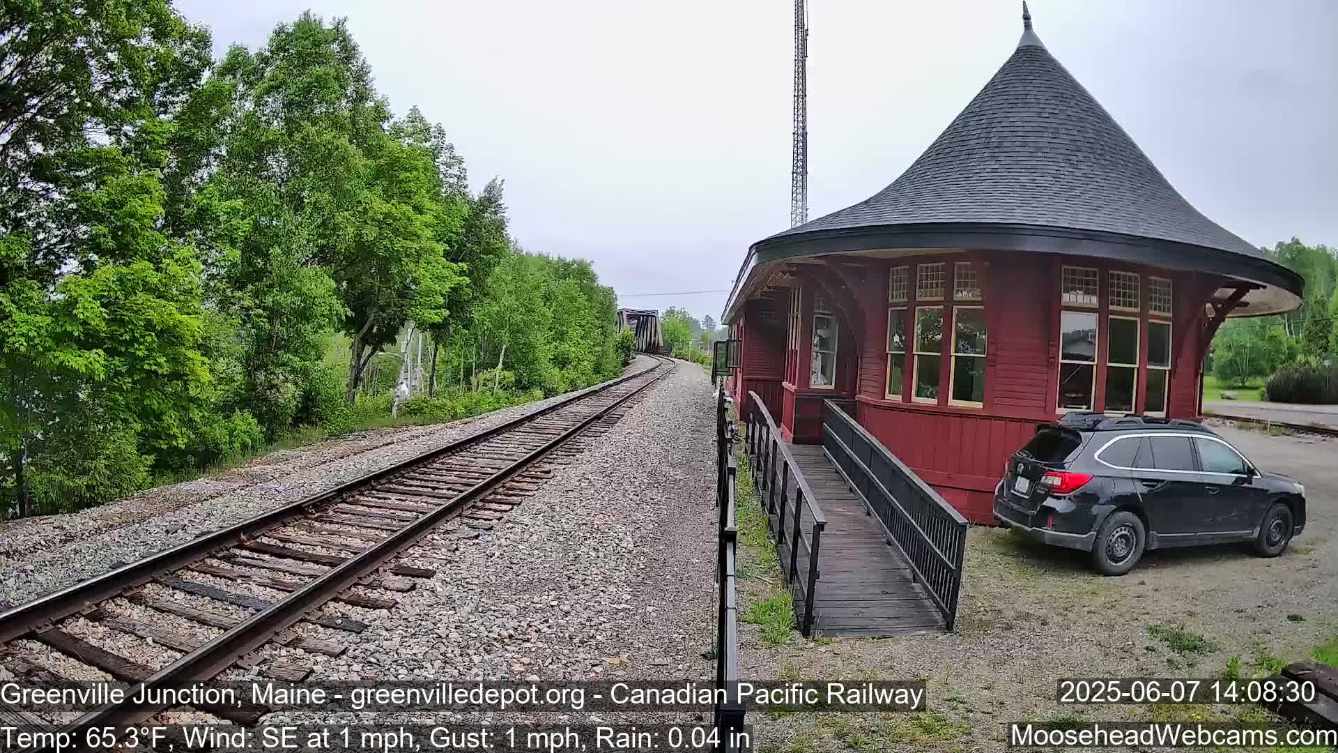 A red, round train station with a dark roof sits next to train tracks that extend into a green, tree-lined landscape under slightly overcast skies with light rain.